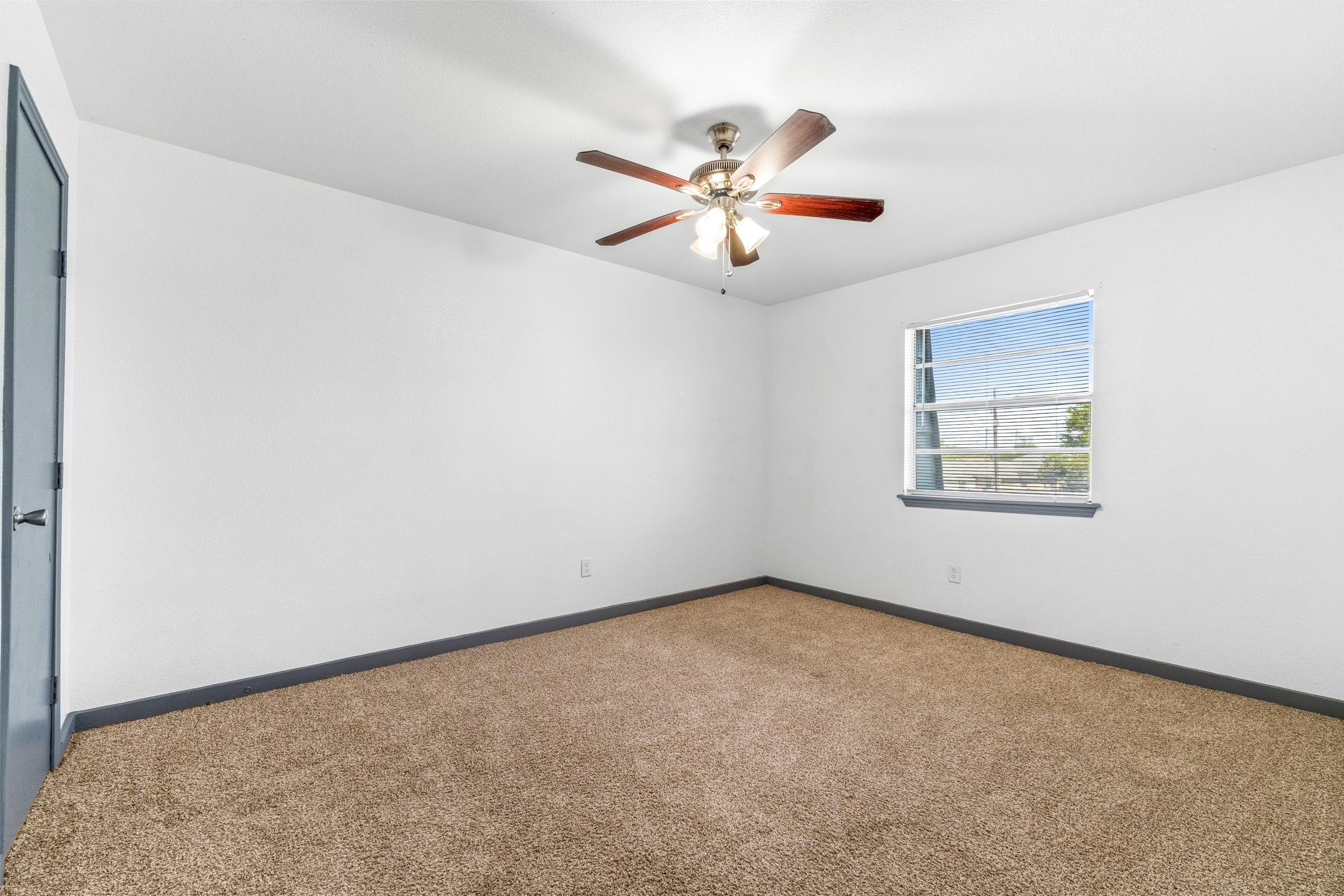 A clean, empty room with light-colored walls and a ceiling fan. The floor is covered in tan carpet, and a window with blinds lets in natural light. There is a blue door on one side, adding a touch of color to the otherwise neutral space. The room appears bright and airy, suitable for various uses.