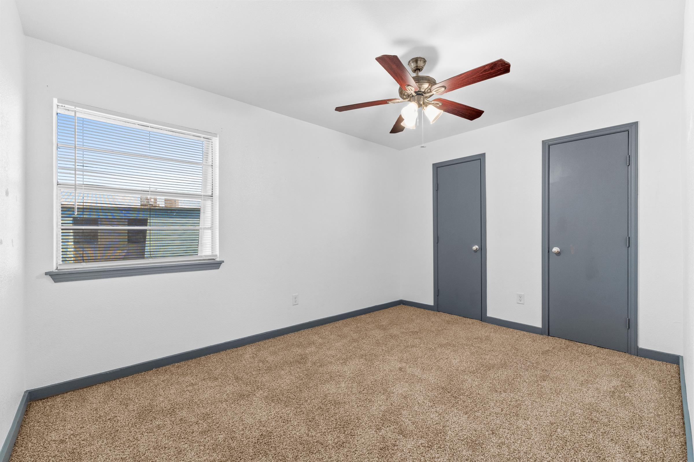 A bright, empty room with beige carpet and white walls. A ceiling fan with wooden blades hangs from the ceiling. There are two closed gray doors on one side and a window with blinds that lets in natural light, offering a view of the outside. The overall ambiance is clean and simple.
