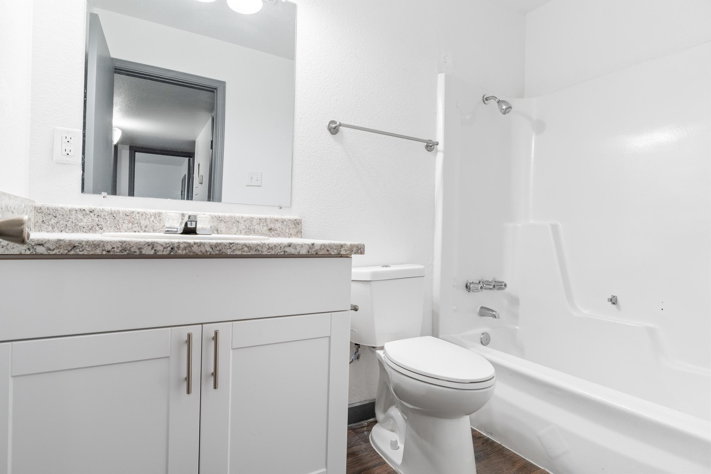 A clean and modern bathroom featuring a white bathtub with a shower, a sink with a granite countertop, a toilet, and light-colored walls. In the background, there is a doorway leading to another room, and the space is well-lit with overhead fixtures.