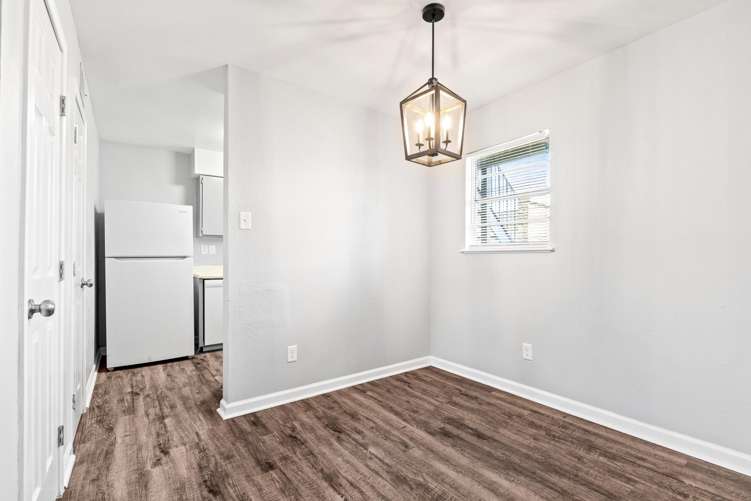A well-lit, empty room featuring light gray walls, a stylish pendant light fixture, and a window. The floor is covered with wood-like laminate. In the corner, there is a white refrigerator, and two doors are visible, suggesting access to other areas in the space.
