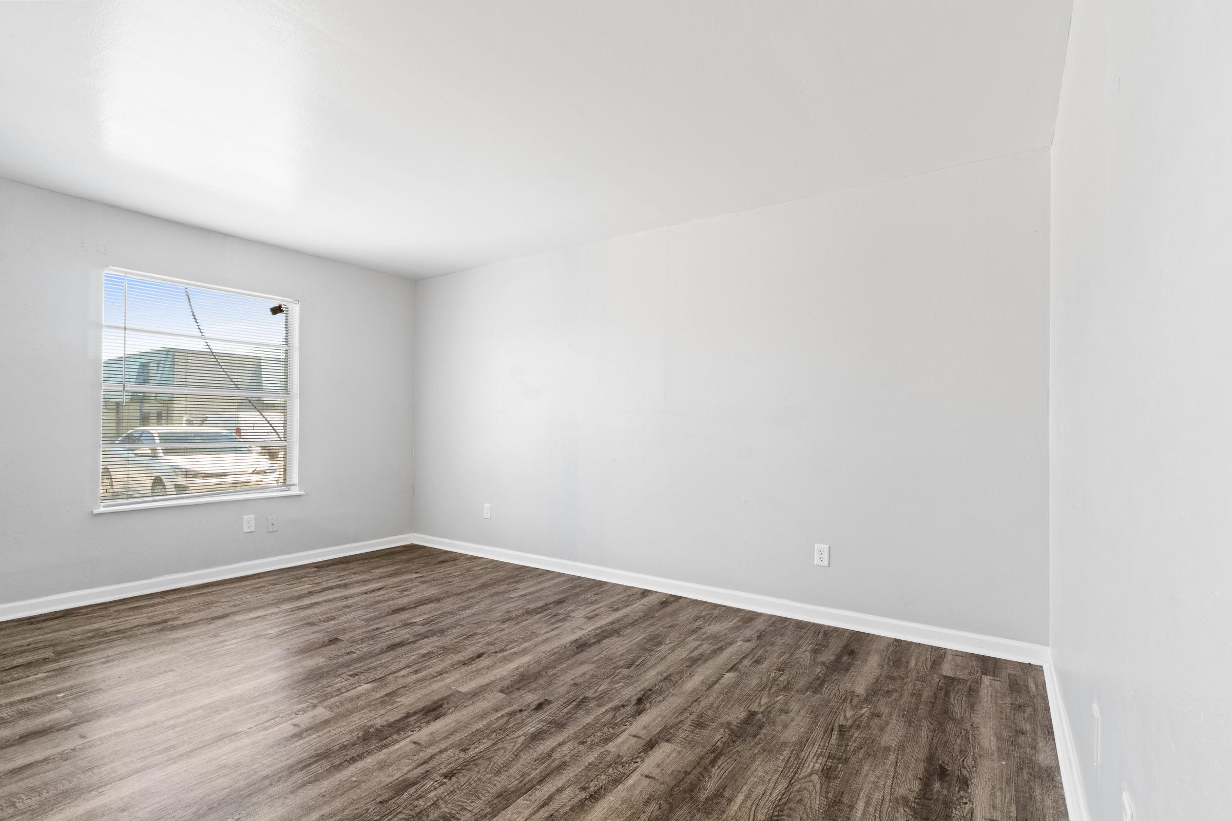 A vacant room with light gray walls and wooden flooring. There's a window on the left side allowing natural light to enter, but the room is otherwise empty with no furniture or decorations. The space has a clean and modern appearance.