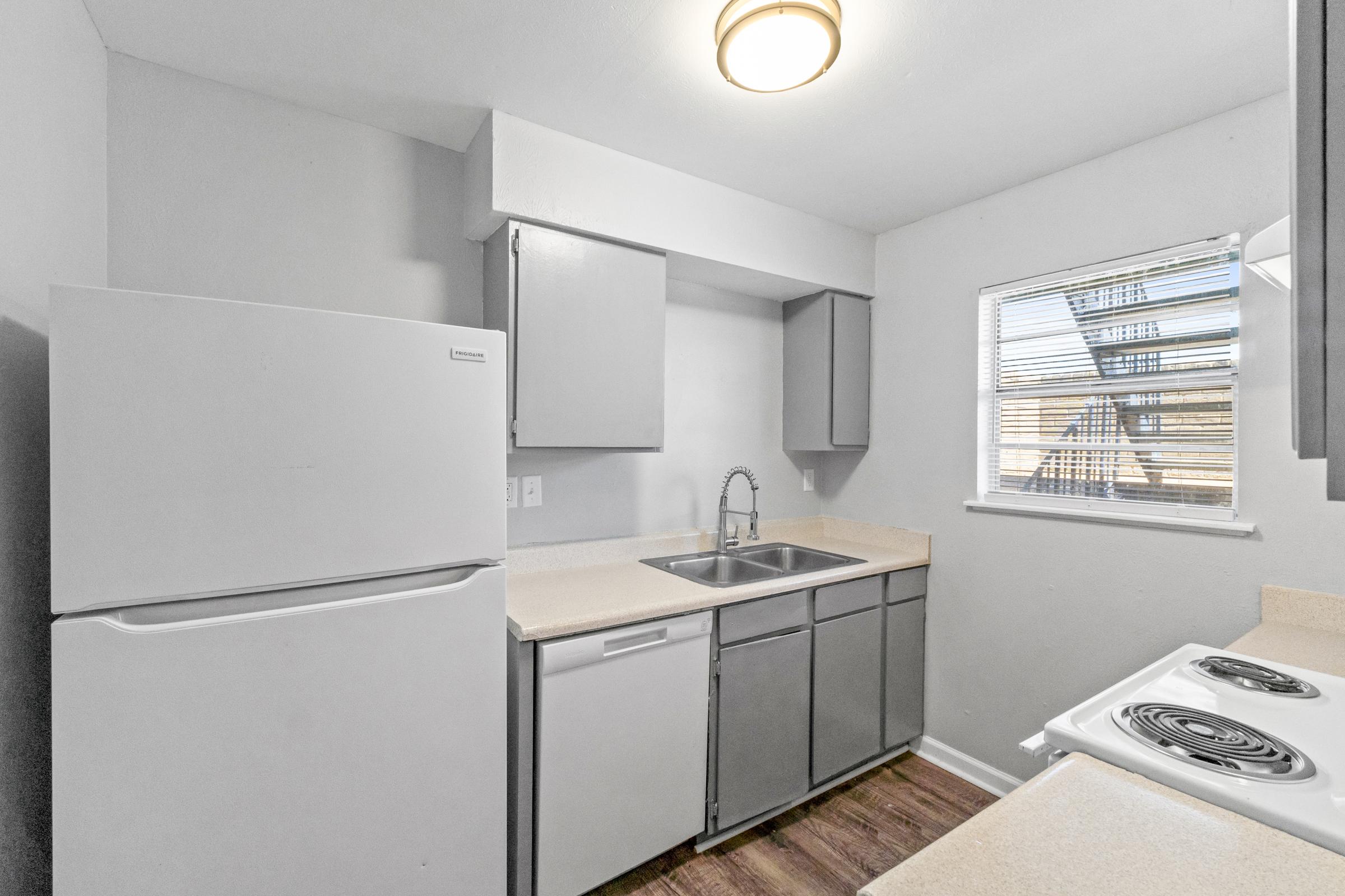 A modern kitchen featuring gray cabinetry, a white refrigerator, a stainless steel sink, and an electric stove. The area has a small window with horizontal blinds allowing natural light. The countertop is light-colored, and the flooring is wooden, creating a clean and inviting space.