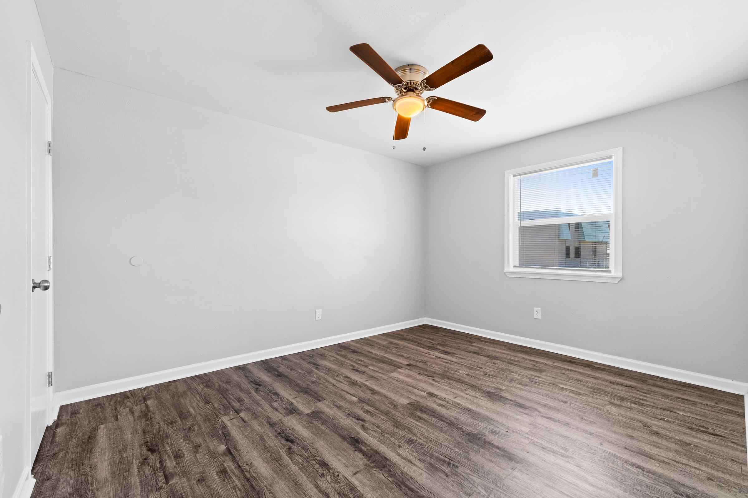 A spacious, empty room with light gray walls and a window on one side. The floor features dark wood-like laminate, and a ceiling fan with a light fixture is mounted in the center. The overall atmosphere is bright and airy, offering potential for decoration.