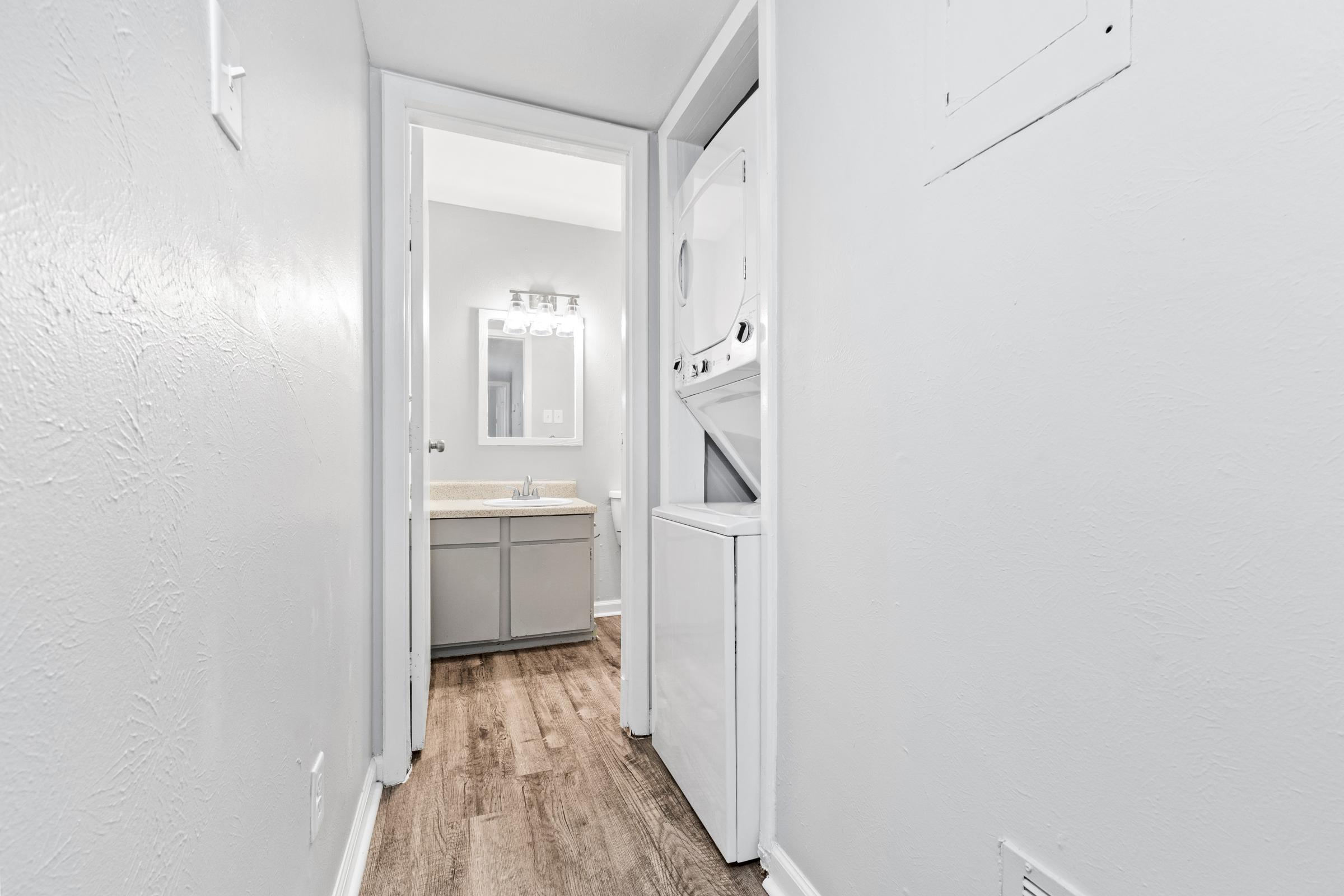 A narrow hallway leading to a bathroom. On the left, a stacked washer and dryer. The bathroom at the end has a vanity with a mirror and bright lighting. The walls are painted light gray, and the floor features wooden planks.