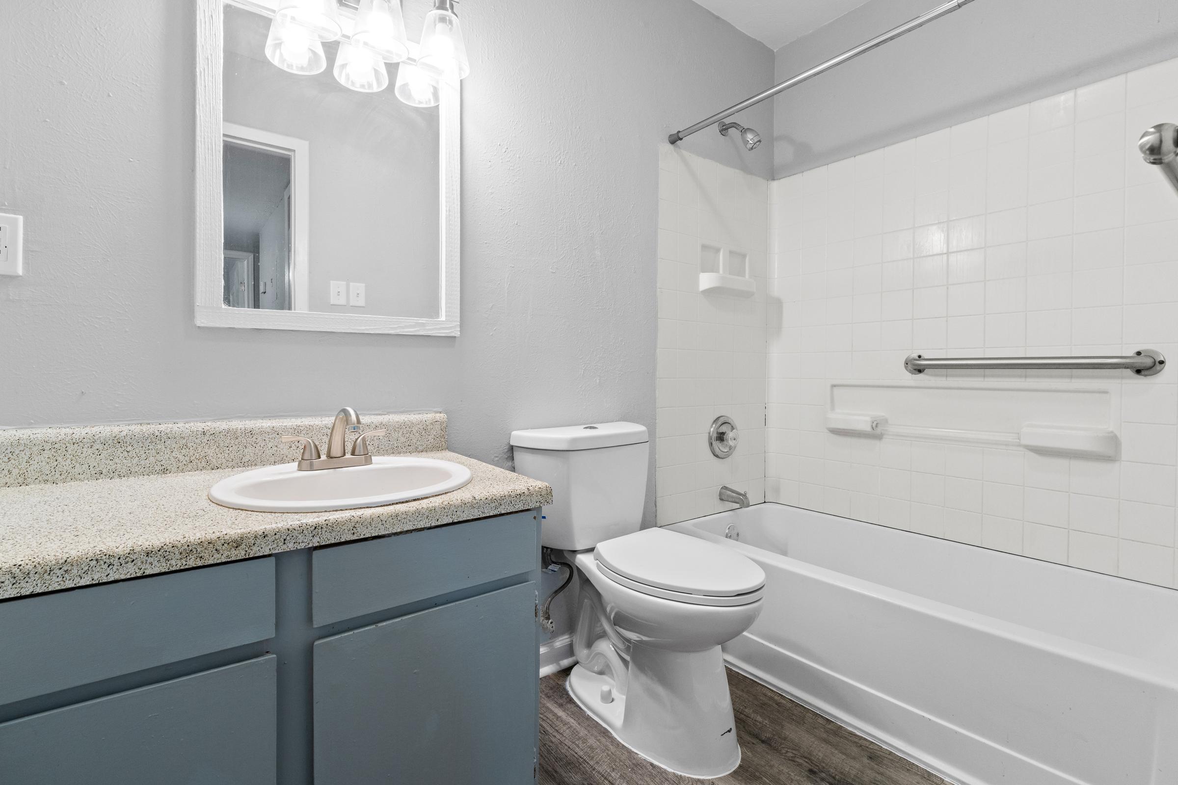 A clean and modern bathroom featuring a white bathtub with a silver grab bar, a toilet, and a sink with a speckled beige countertop. The walls are painted light gray, and there is a mirror above the sink with three lights above it. The flooring appears to be wood-like laminate.