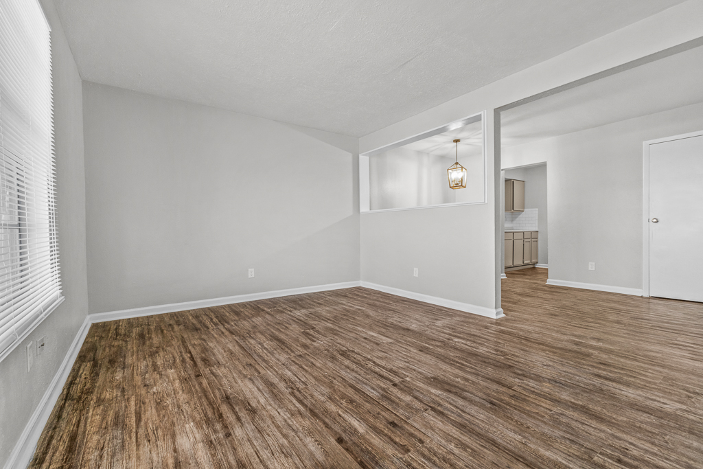 A spacious, newly renovated living area with light-colored walls and hardwood-style flooring. A window with blinds allows natural light, and a doorway leads to a kitchen area in the background, featuring a pendant light and modern cabinetry. The overall ambiance is bright and inviting.