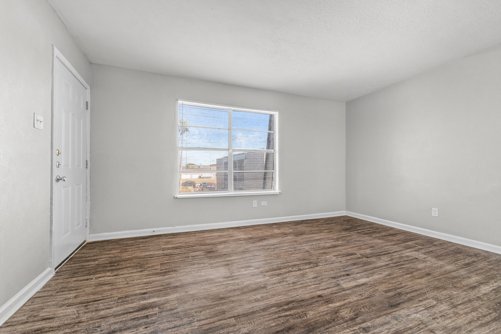 A spacious, empty room with light gray walls and a window allowing natural light. The floor is a light-colored wood laminate. There's a white door on the left and minimal decor, creating a clean and modern feel.