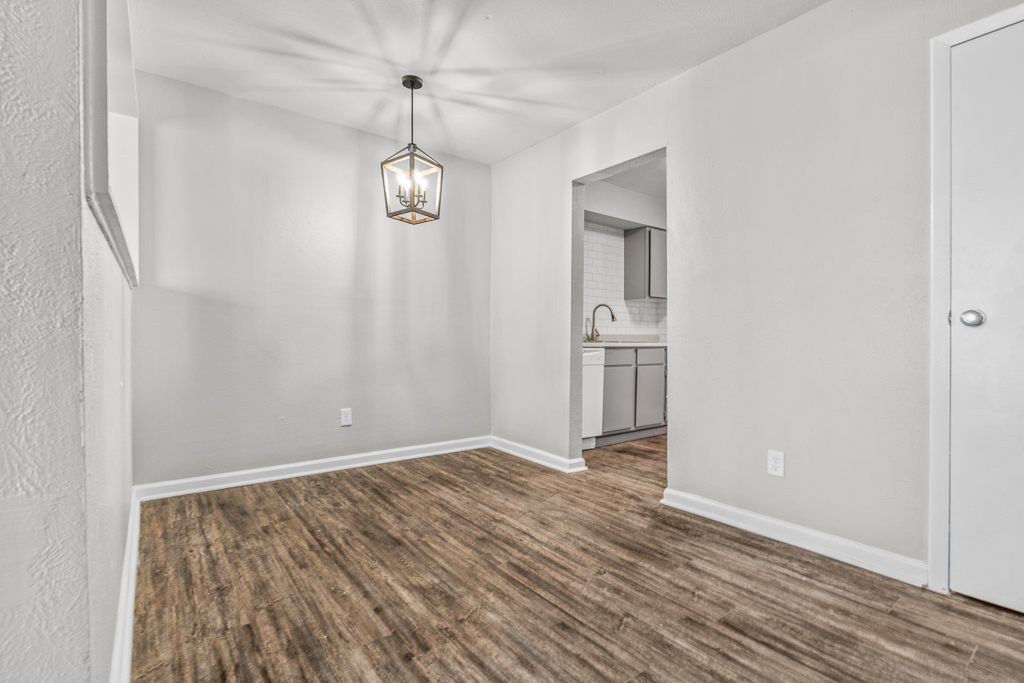 A well-lit interior space featuring light-colored walls and a modern pendant light. The room has a wood-style floor and is partially open to a kitchen area visible through a rectangular opening. A white door is seen on the right, complementing the clean and minimalist design of the room.