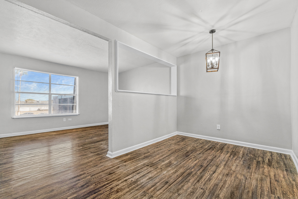 Bright, spacious interior of a living room featuring wooden flooring, a large window allowing natural light, and a contemporary hanging light fixture. The walls are painted in a light color, creating an airy atmosphere. A mirrored section is present, adding depth to the space.