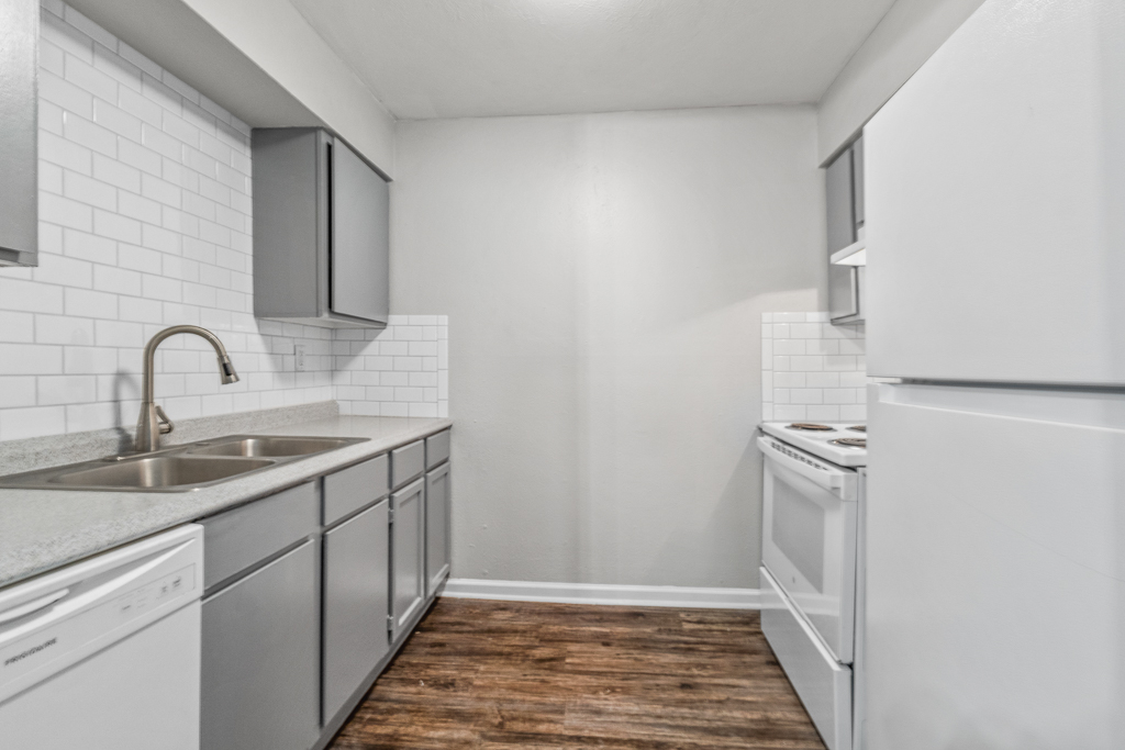 Modern kitchen featuring gray cabinets, white subway tile backsplash, stainless steel sink, dishwasher, and a white refrigerator. The space has a clean, minimalist design with wood laminate flooring and ample natural light.