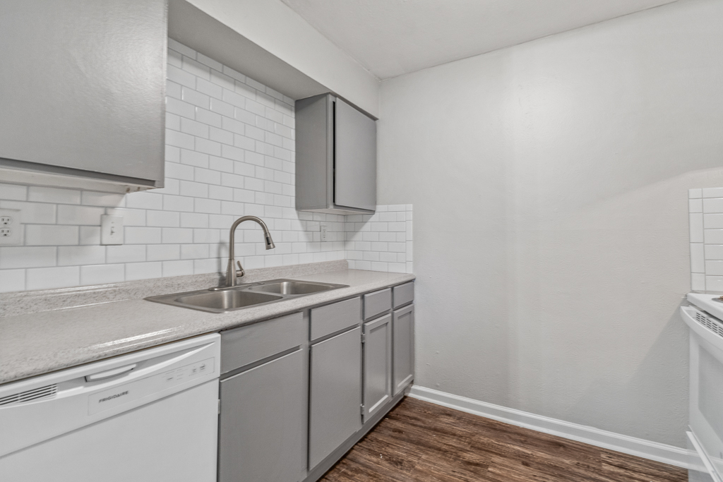 A modern kitchen featuring grey cabinets, a double sink with a sleek faucet, white subway tile backsplash, and a dishwasher. The flooring is wood-like, and the kitchen is well-lit with a neutral-colored wall providing a clean and spacious look.