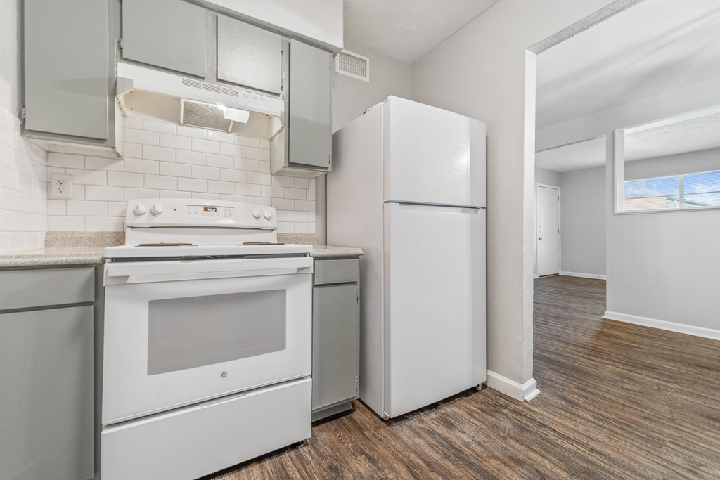 A modern kitchen featuring a white stove, hood, and refrigerator with gray cabinetry. The walls are painted light gray, and the floor has a wood-like finish. A doorway leads to a bright, open living space with windows visible in the background.
