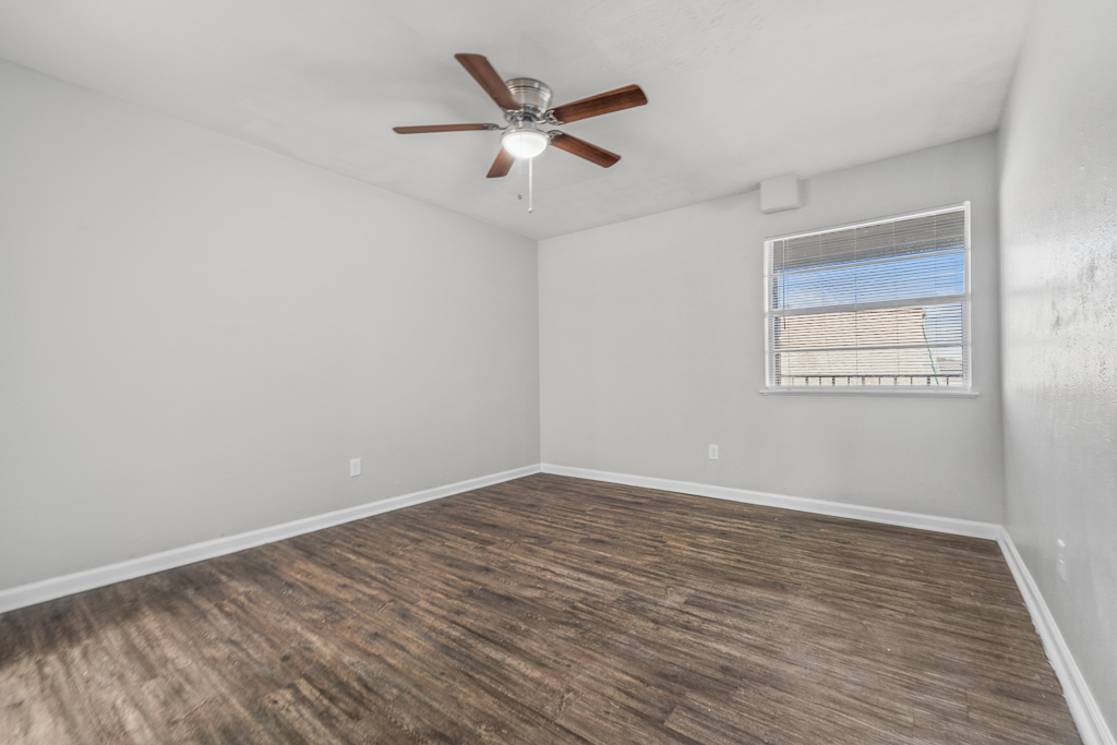 Empty room with gray walls and a ceiling fan. The floor is made of wood-like laminate, and there is a window with blinds letting in natural light. The room appears clean and well-maintained, suitable for various uses.