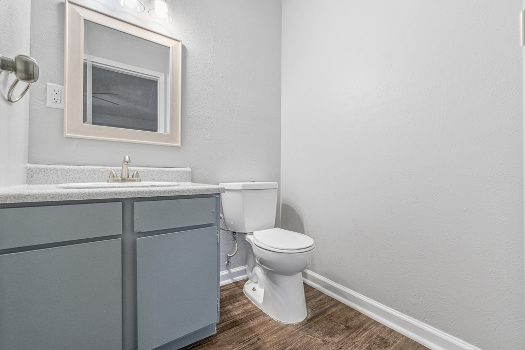 A clean, modern bathroom featuring a light gray wall, a simple wooden mirror above the sink, a countertop with a faucet, and a white toilet. The floor is laminate with a wood-like finish, creating a tidy and minimalist aesthetic.