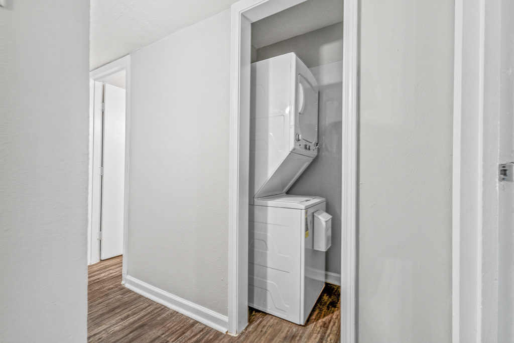 A narrow hallway leading to a closet featuring a stacked washer and dryer. The walls are painted light gray, and the flooring is a wood-like material. The scene is well-lit, emphasizing the clean and modern look of the laundry area.