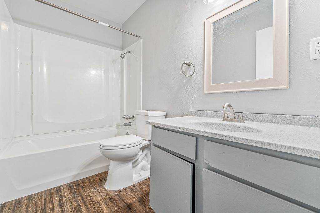 A modern bathroom featuring a white bathtub and shower combination, a toilet, and a single sink vanity with a grey countertop. The walls are painted in a light grey color, and there is a simple mirror above the sink. The flooring is a wood-like laminate.