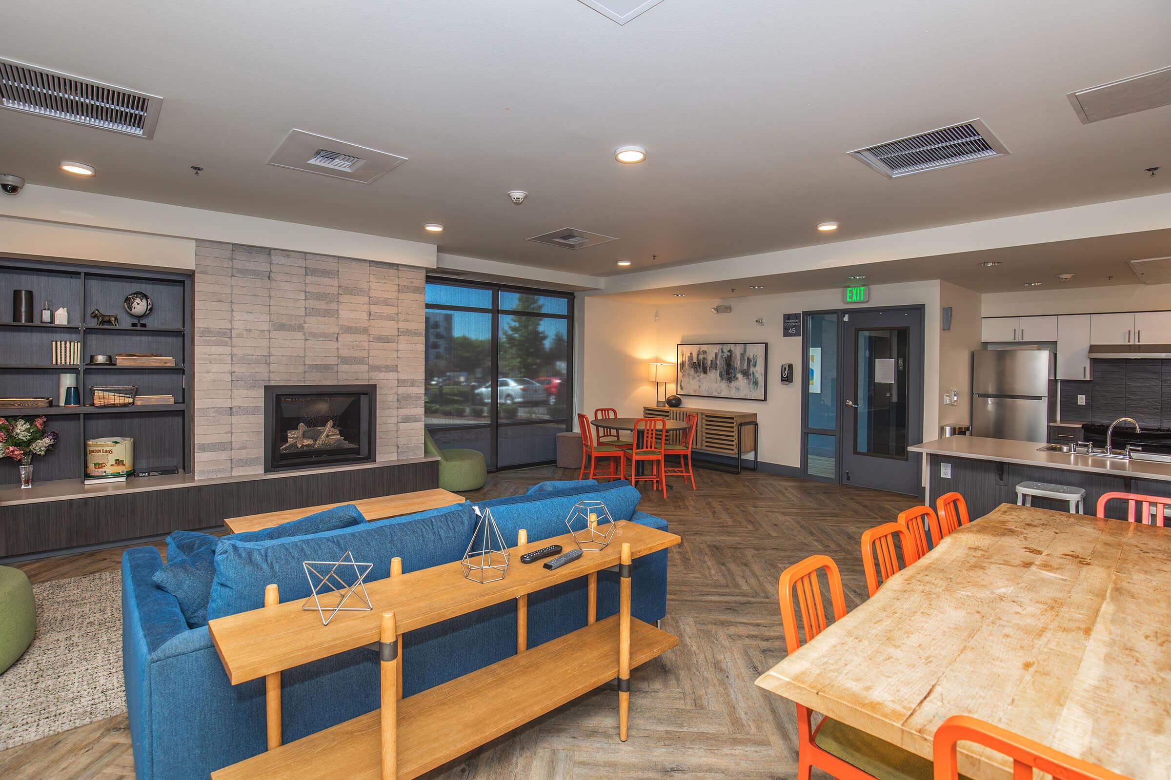 A cozy lounge area featuring a blue sofa, wooden dining table with orange chairs, and a modern fireplace. The room has large windows letting in natural light, with a bookshelf and decorative items enhancing the inviting atmosphere. Ideal for relaxation and socializing.