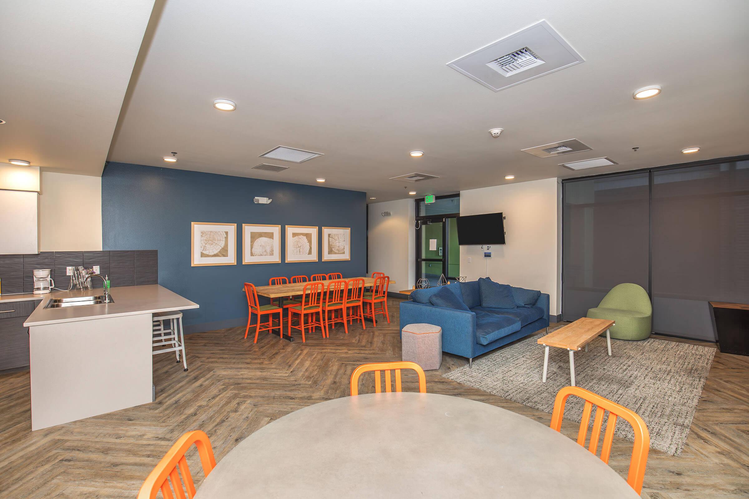 A modern, open living space featuring a cozy blue sofa, a wooden coffee table, and orange dining chairs around a large table. The walls are painted in soft tones, with framed artwork displayed. A kitchenette is visible in the background, and large windows let in natural light.