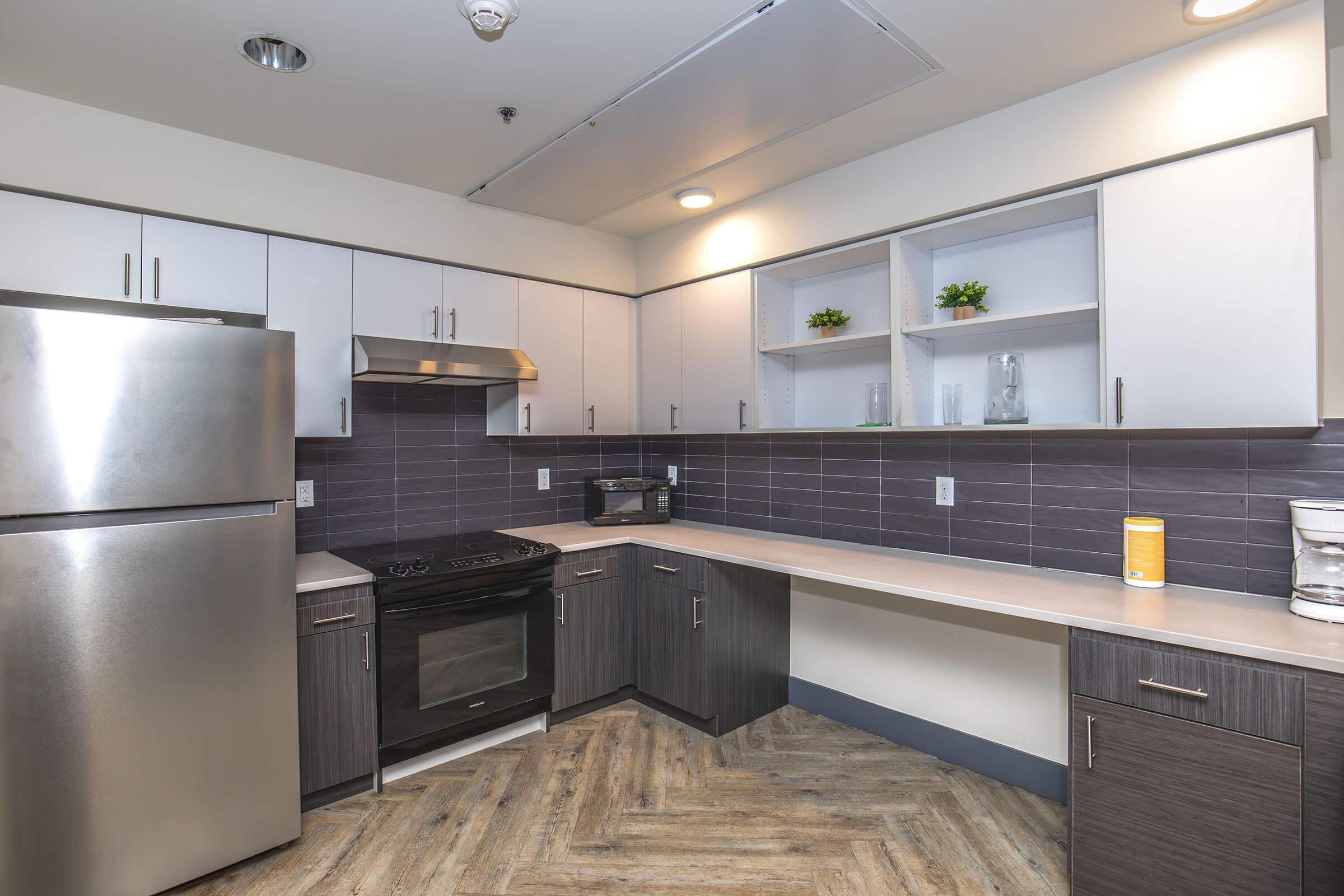 A modern kitchen featuring stainless steel appliances, including a refrigerator, stove, and microwave. The cabinetry has a sleek design with gray and white finishes. Open shelves are displayed with decorative plants, while the countertops are light-colored, and the flooring has a wood-like appearance.