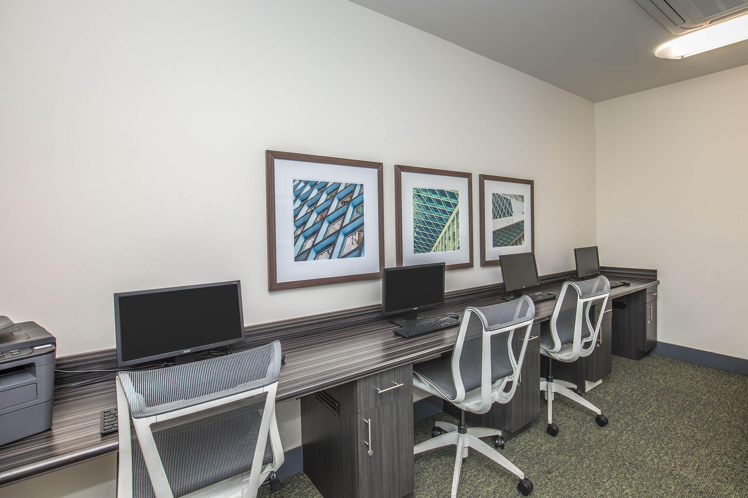 A modern workspace featuring four computer stations with ergonomic chairs, a printer, and a simple wooden desk. The wall is adorned with four framed images of architectural details. The room has neutral colors and a clean, organized appearance, ideal for productivity and focusing on tasks.