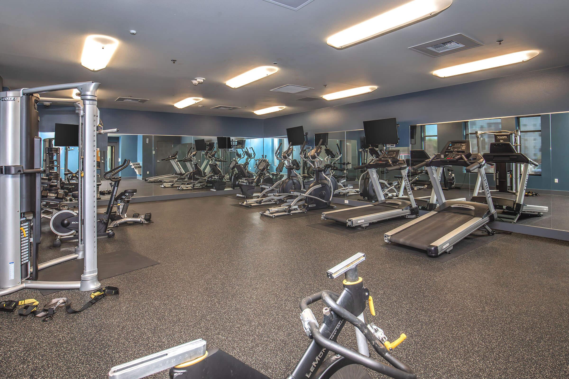A spacious gym featuring a variety of exercise equipment, including treadmills, elliptical machines, and weight machines. The room has large mirrors and fluorescent lighting, creating a bright and inviting atmosphere for workouts. A clean and modern design is evident throughout the space.
