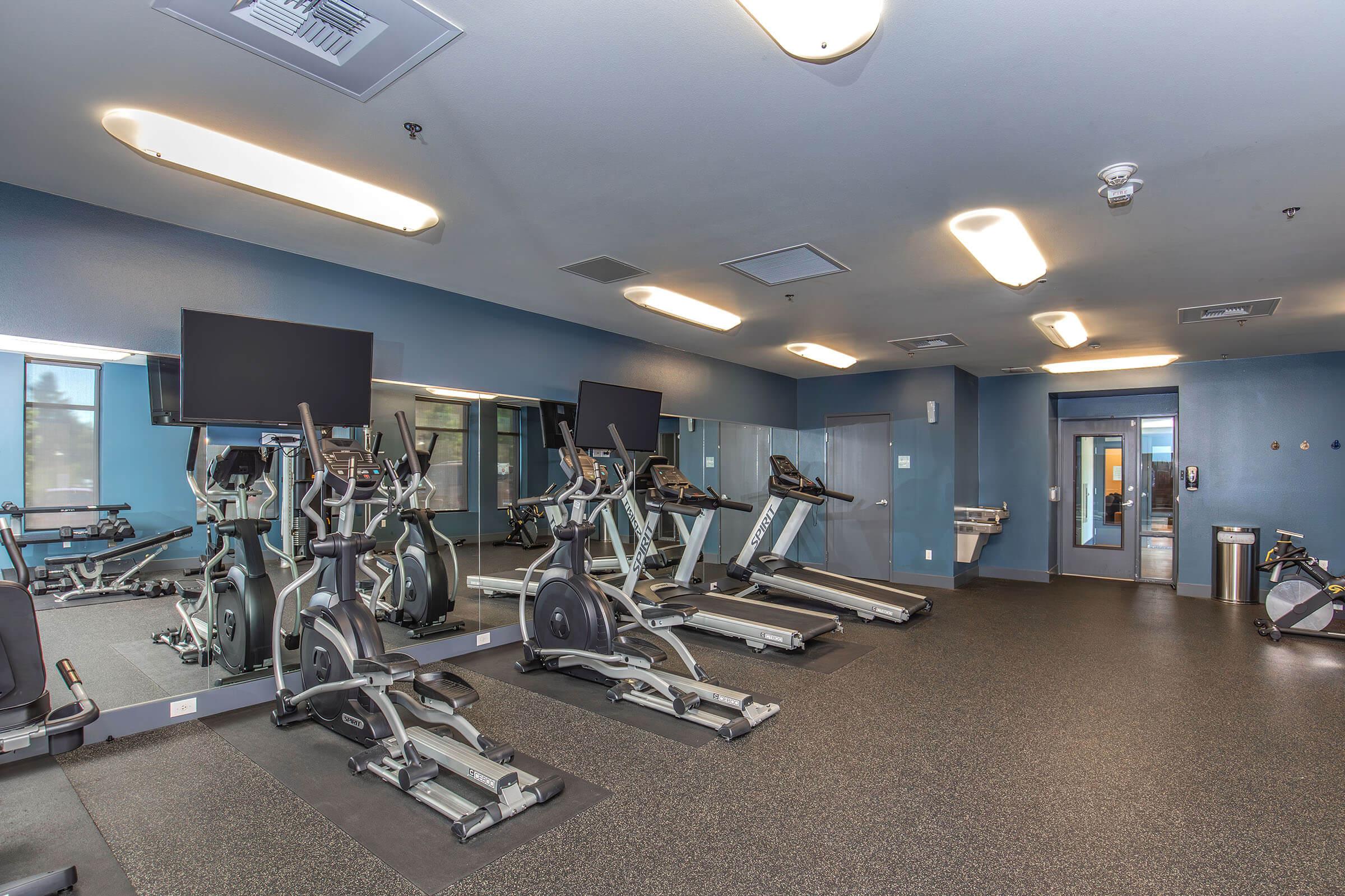 A modern gym interior featuring exercise equipment, including treadmills and ellipticals, positioned on a rubber floor. There are large mirrors on one wall and flat-screen TVs mounted above the machines. The space is well-lit with overhead lights and has a clean, contemporary design.