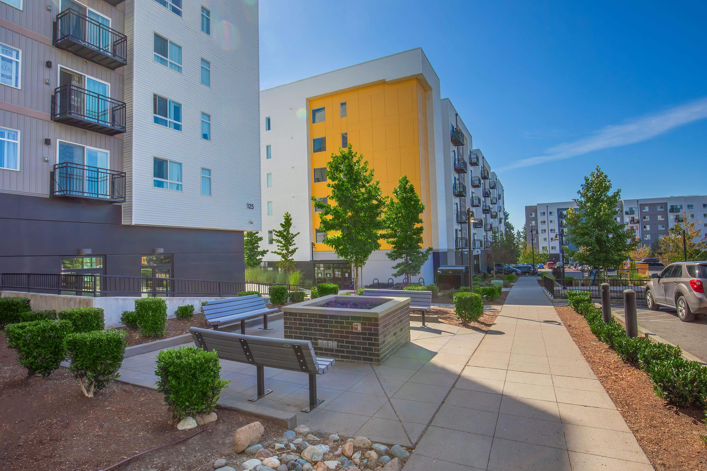 A pathway lined with small shrubs and benches leads through a modern residential area. On one side, there is a fire pit. The buildings feature a mix of textures and colors, with one prominently displaying a bright yellow facade under a clear blue sky.