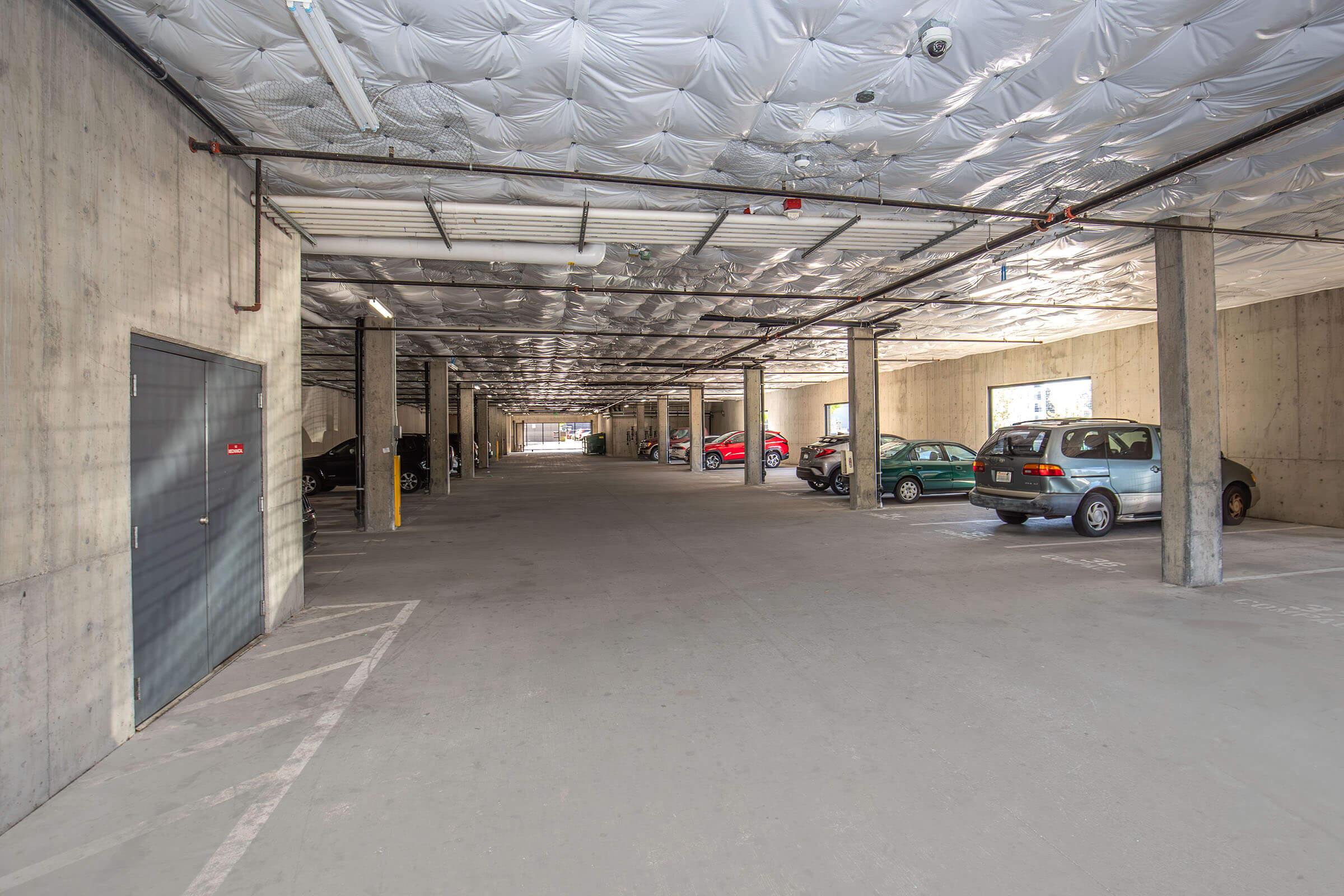 A spacious underground parking garage featuring concrete walls and a ceiling with a unique texture. Several parked cars are visible, including a green vehicle and a grey minivan. The area is well-lit, with parking spaces marked on the ground and an entrance visible in the distance.
