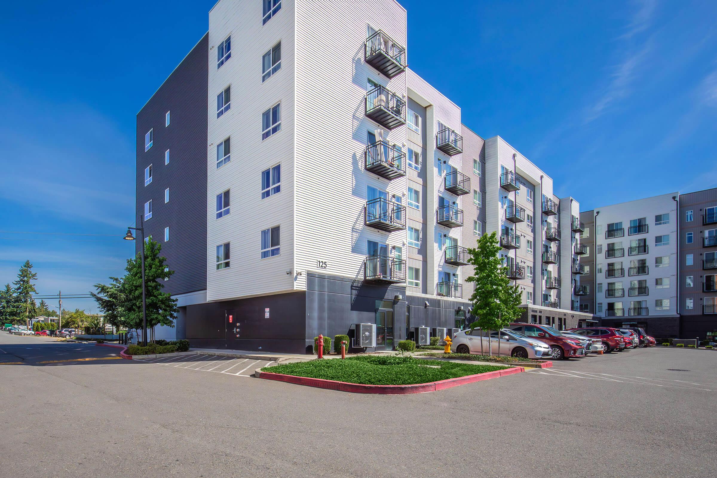 A modern multi-story apartment building with a mix of balconies and large windows, surrounded by a parking lot. The scene features clear blue skies and well-maintained landscaping, including green trees and shrubs, with several parked cars in the foreground.