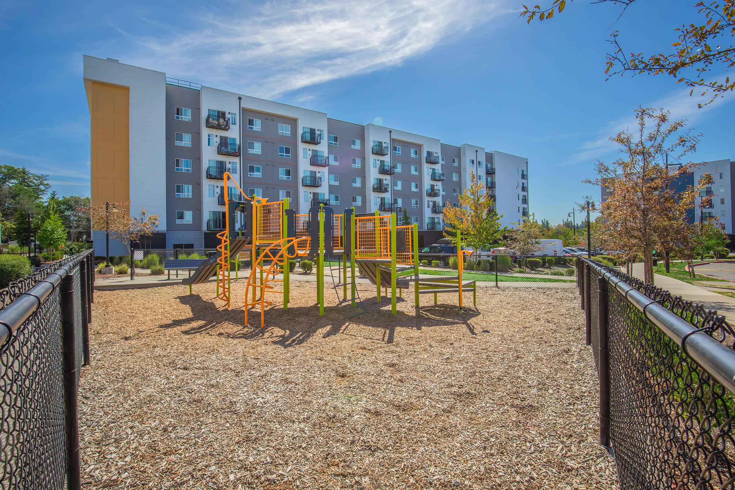 A sunny park featuring a playground with orange climbing structures surrounded by wood chips. In the background, there are modern apartment buildings with balconies. Trees with autumn foliage add greenery to the scene under a clear blue sky.