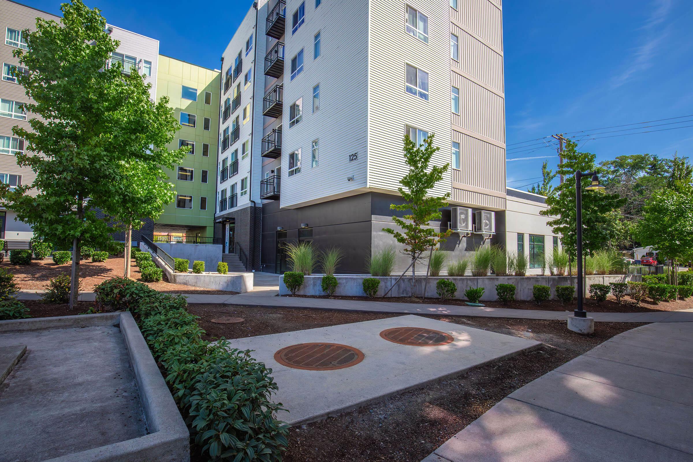 A modern apartment building with multiple stories surrounded by landscaped gardens and greenery. The foreground features a paved area with decorative elements, while the background showcases well-maintained bushes and trees against a clear blue sky.