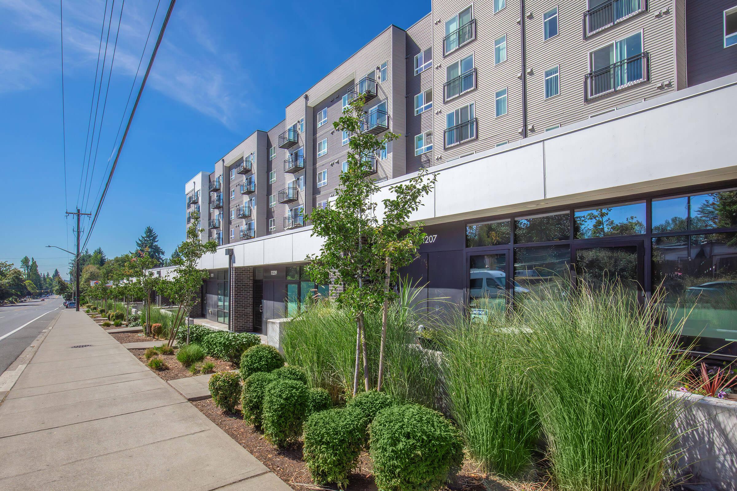 Modern building with multiple stories featuring rectangular windows and balconies, surrounded by landscaped greenery including shrubs and ornamental grass. A sidewalk runs along the front, with utility poles in the background and a clear blue sky above.