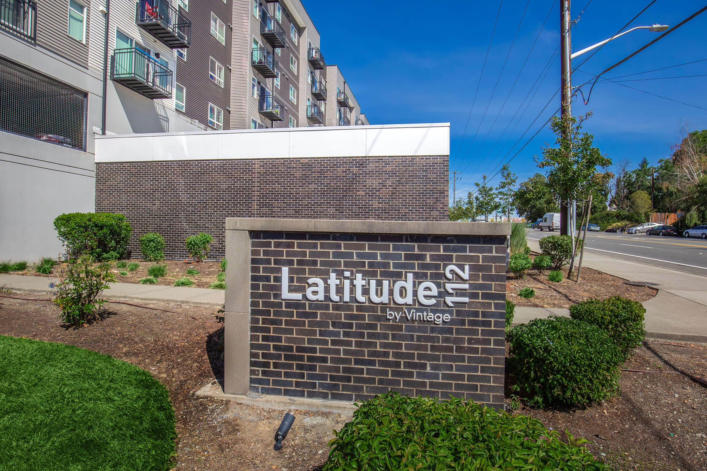 Signage for Latitude 112 by Vintage, featuring modern architecture with nearby greenery and a clear blue sky. The building is seen behind the sign, and the area has well-maintained landscaping.