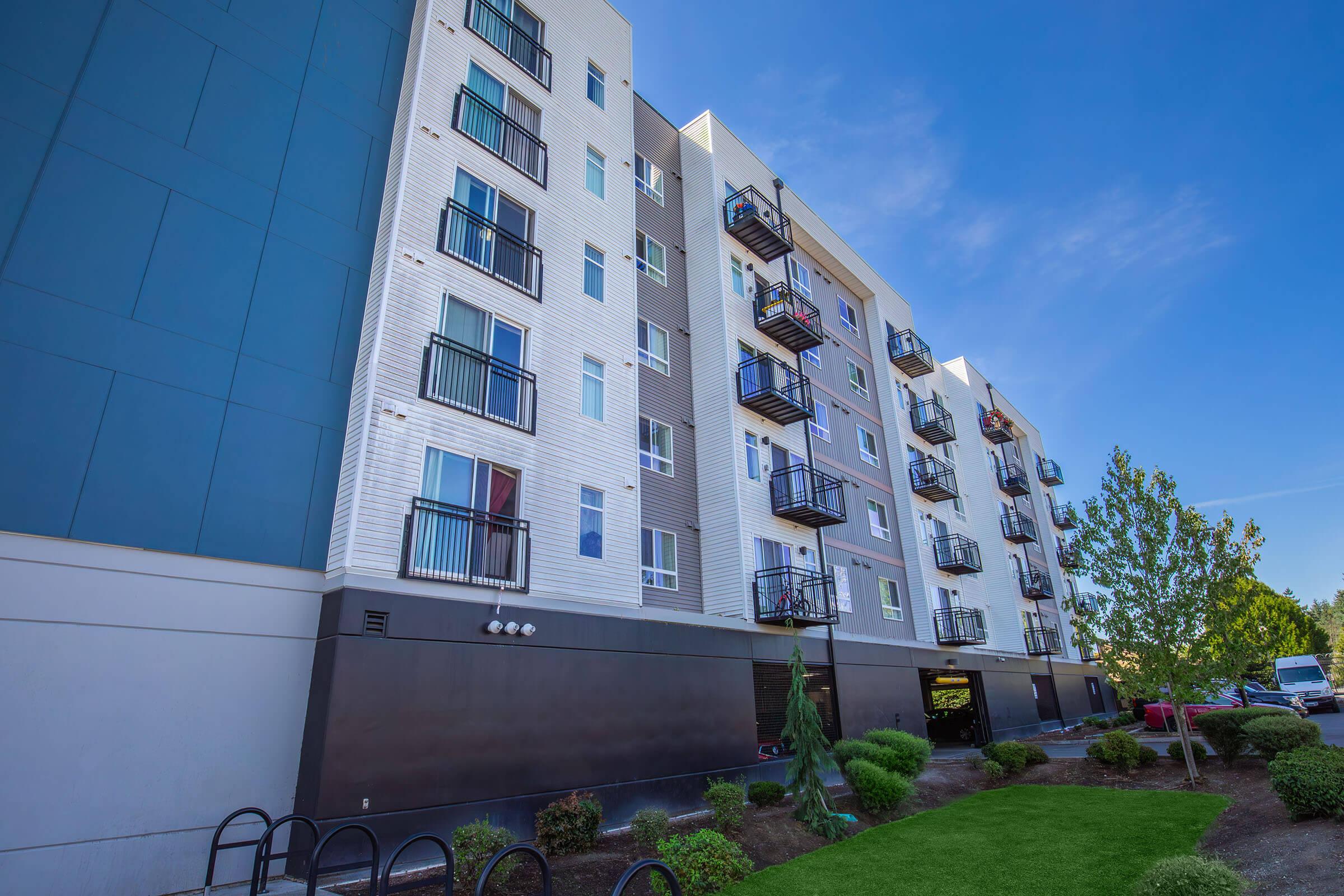 A modern multi-story apartment building with a combination of white and dark exterior. The structure features several balconies, surrounded by well-maintained landscaping and a green lawn. Clear blue sky overhead enhances the vibrant and inviting atmosphere of the setting.