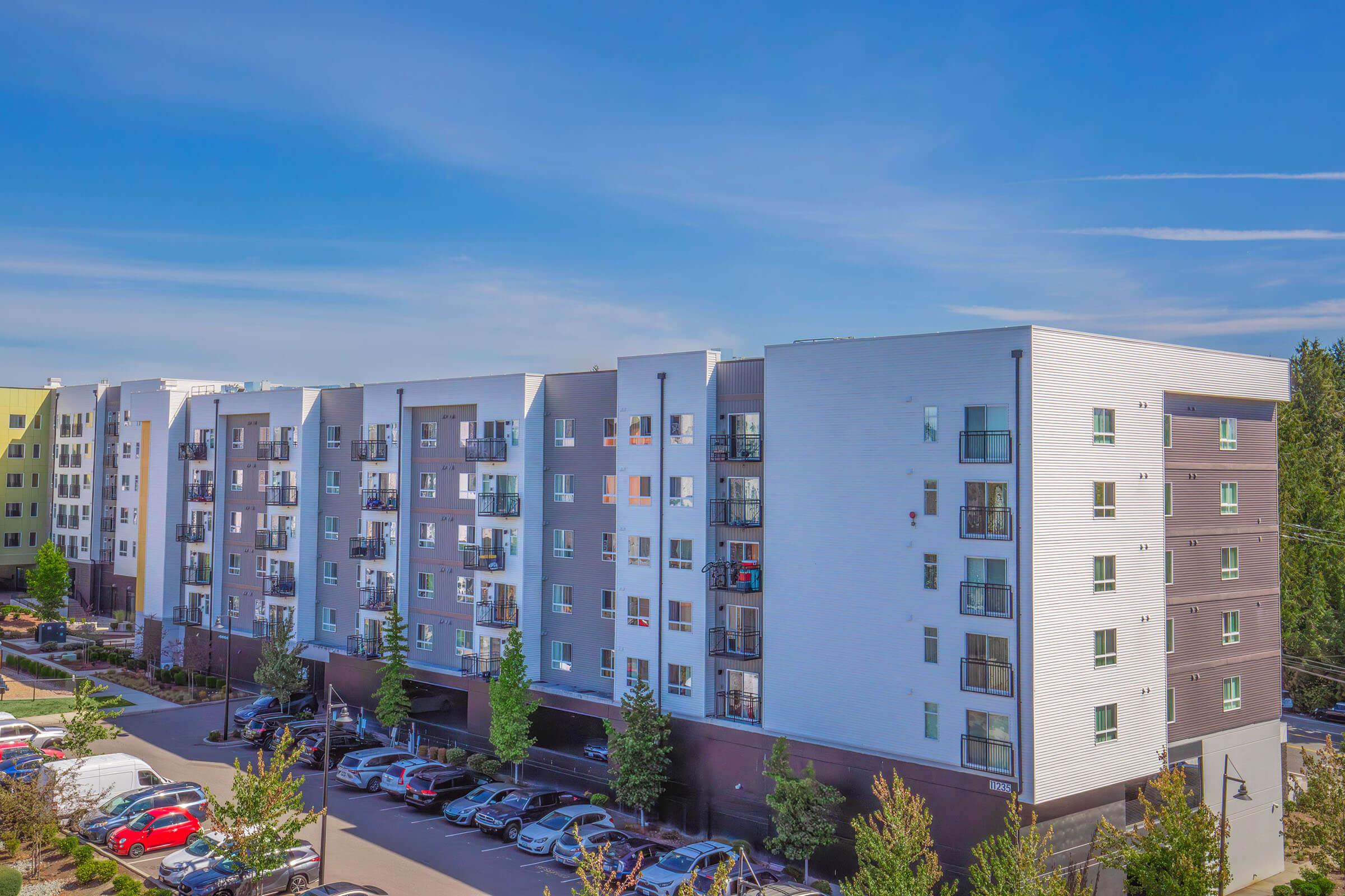 A modern multi-story apartment building with a light and dark exterior, featuring balconies and landscaped surroundings. The parking area in front is filled with various parked cars, and clear blue skies are visible above.