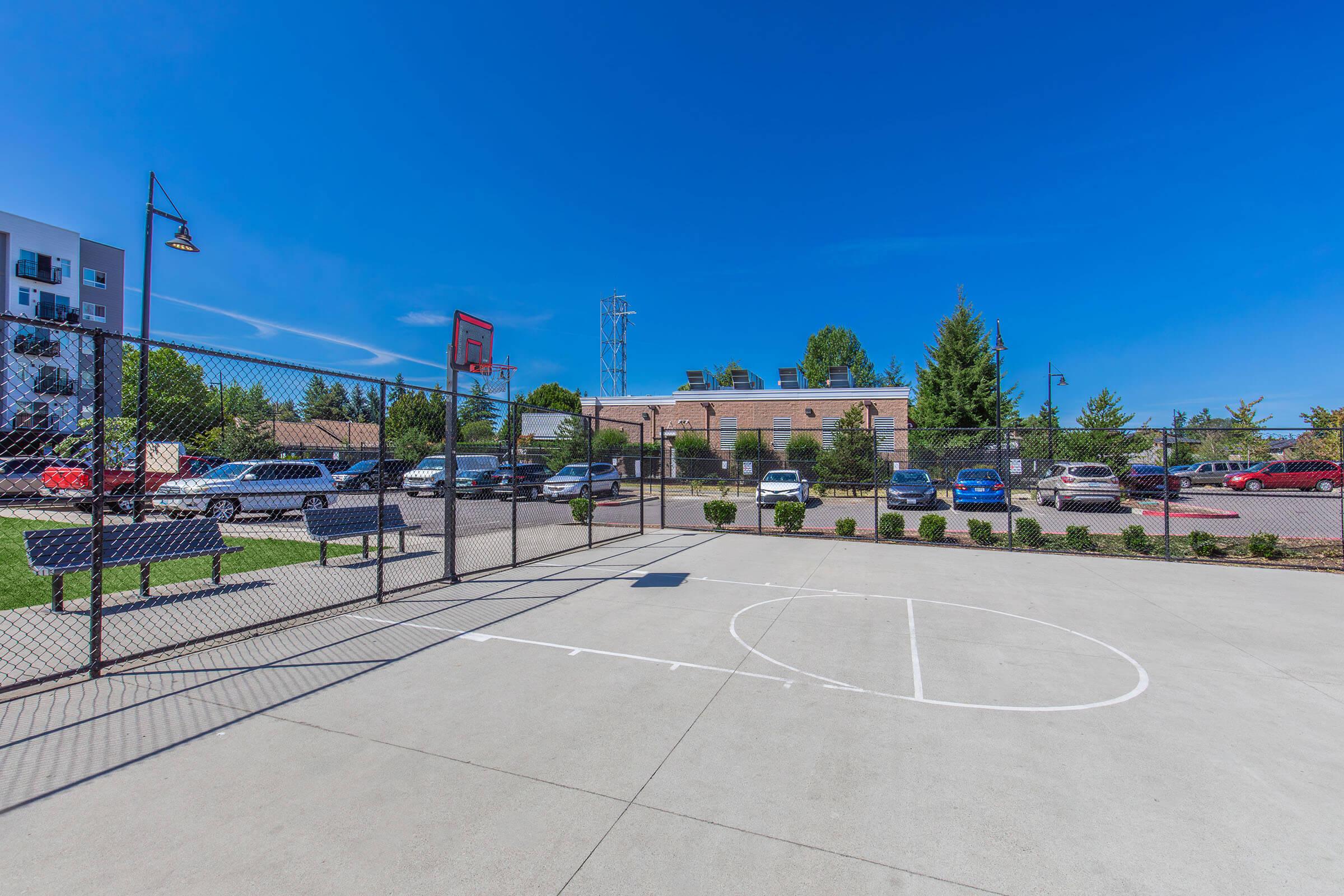A sunny outdoor basketball court enclosed by a fence, featuring a hoop at one end. The court has clear markings, and in the background, there are parked cars and trees. Blue skies and a few clouds are visible above, creating a bright and inviting atmosphere.