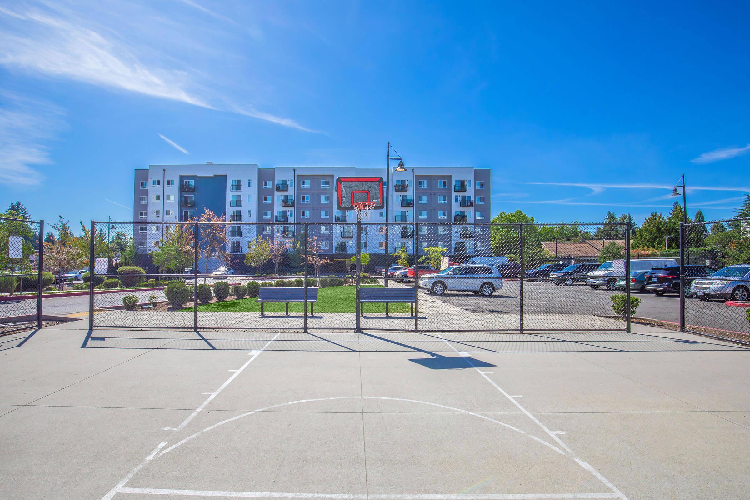 A basketball court with a hoop in the foreground, facing a modern apartment building in the background. The sky is clear and blue, and there are trees and landscaping around the court, with a parking area visible behind the building.