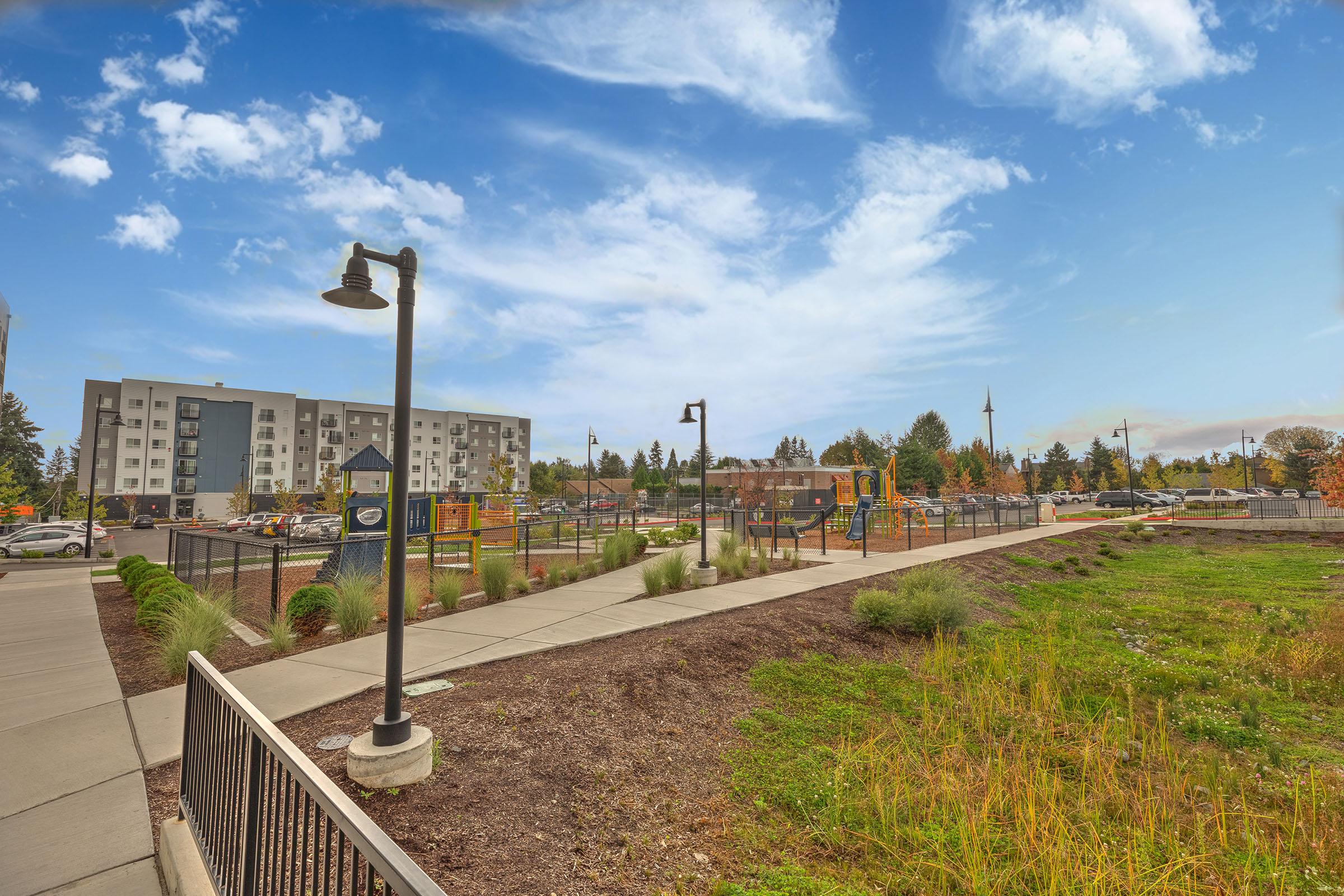 A pathway alongside a green area leading to a playground, with modern buildings in the background under a partly cloudy sky.