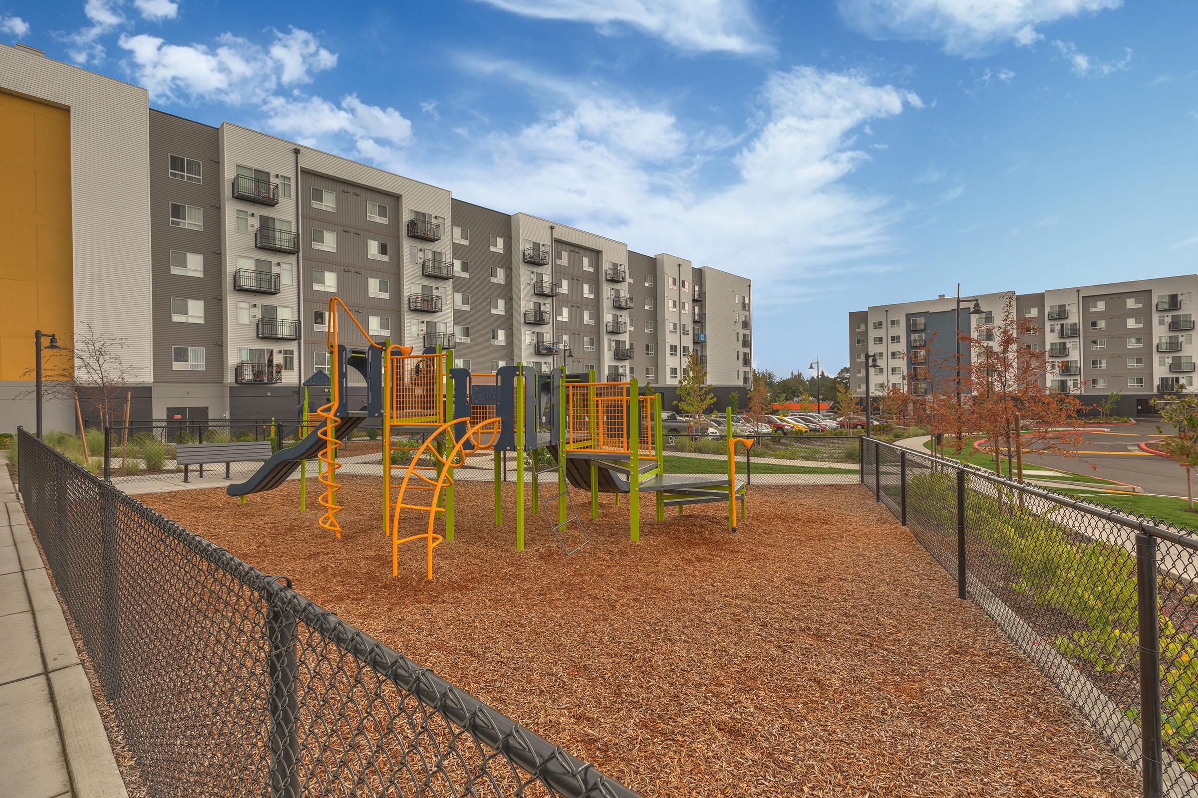 Playground area featuring a colorful play structure with slides and climbing equipment, surrounded by wood chips, alongside modern apartment buildings and a clear blue sky.