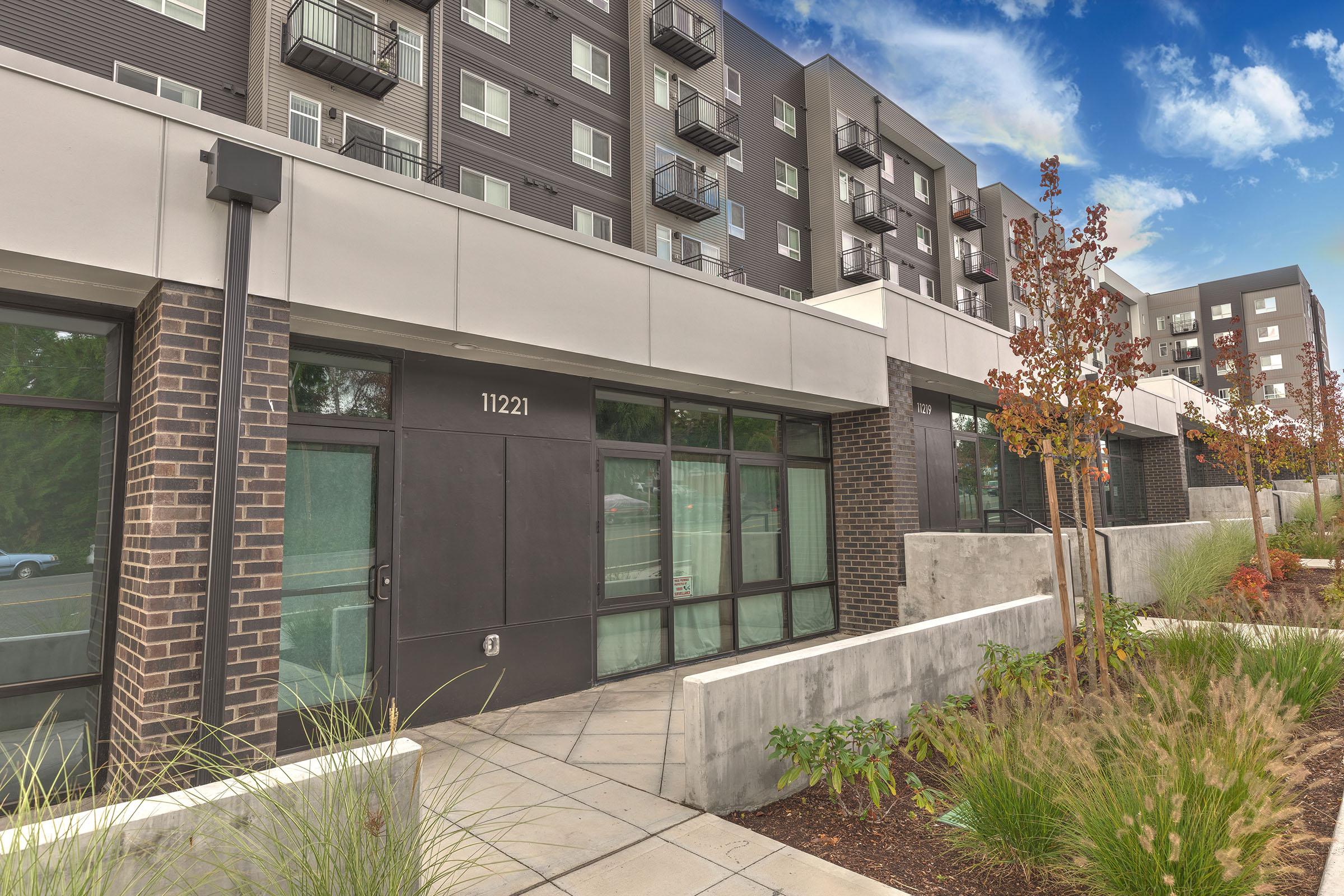 Contemporary apartment building exterior with large windows, black balconies, and landscaped entryway, set against a blue sky.