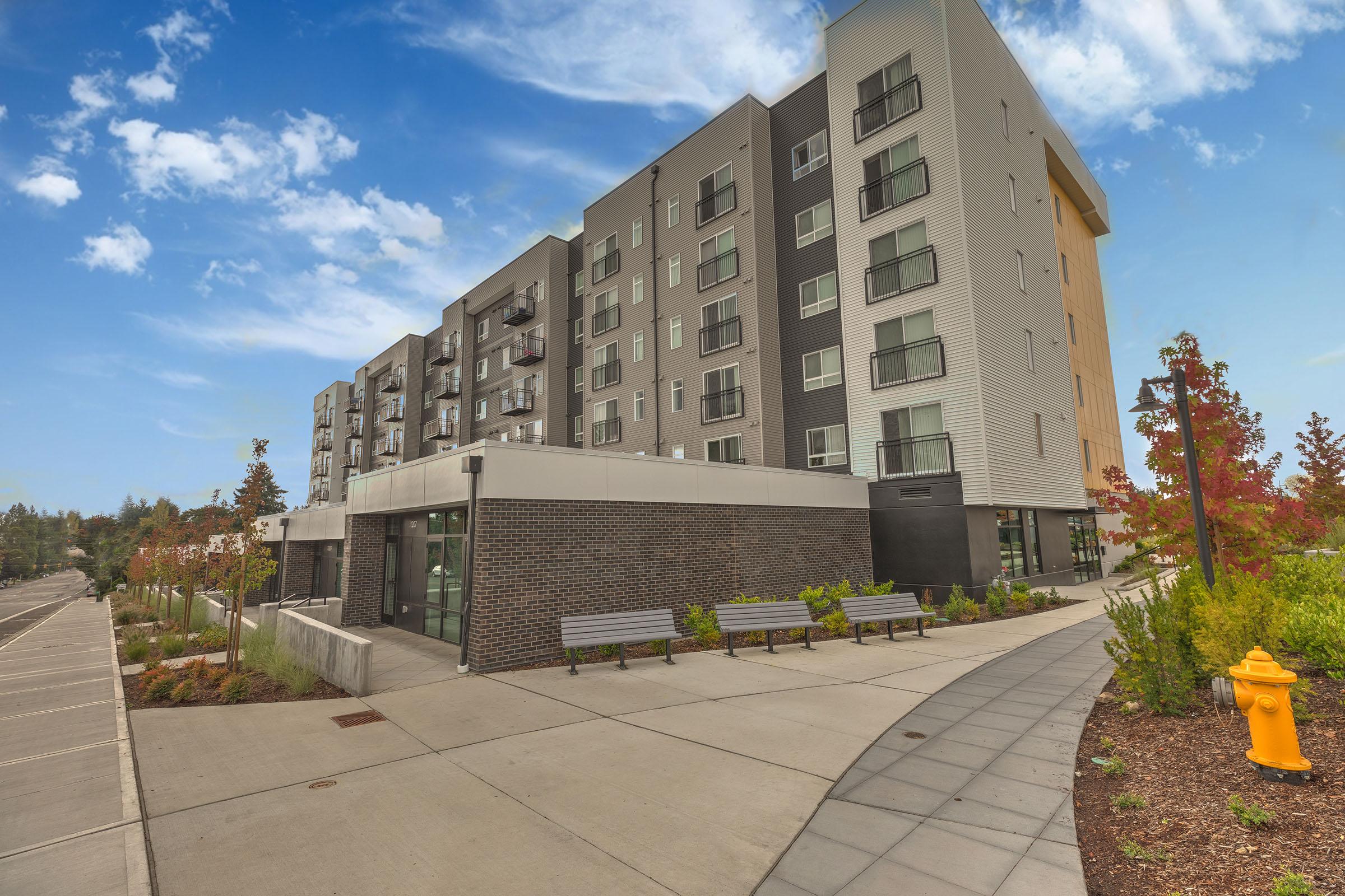 A modern multi-story apartment building with a mix of gray and white exterior, featuring balconies and large windows. Surrounding the building are landscaped areas with benches, trees, and a fire hydrant. Clear blue sky above and a paved walkway leading towards the building.