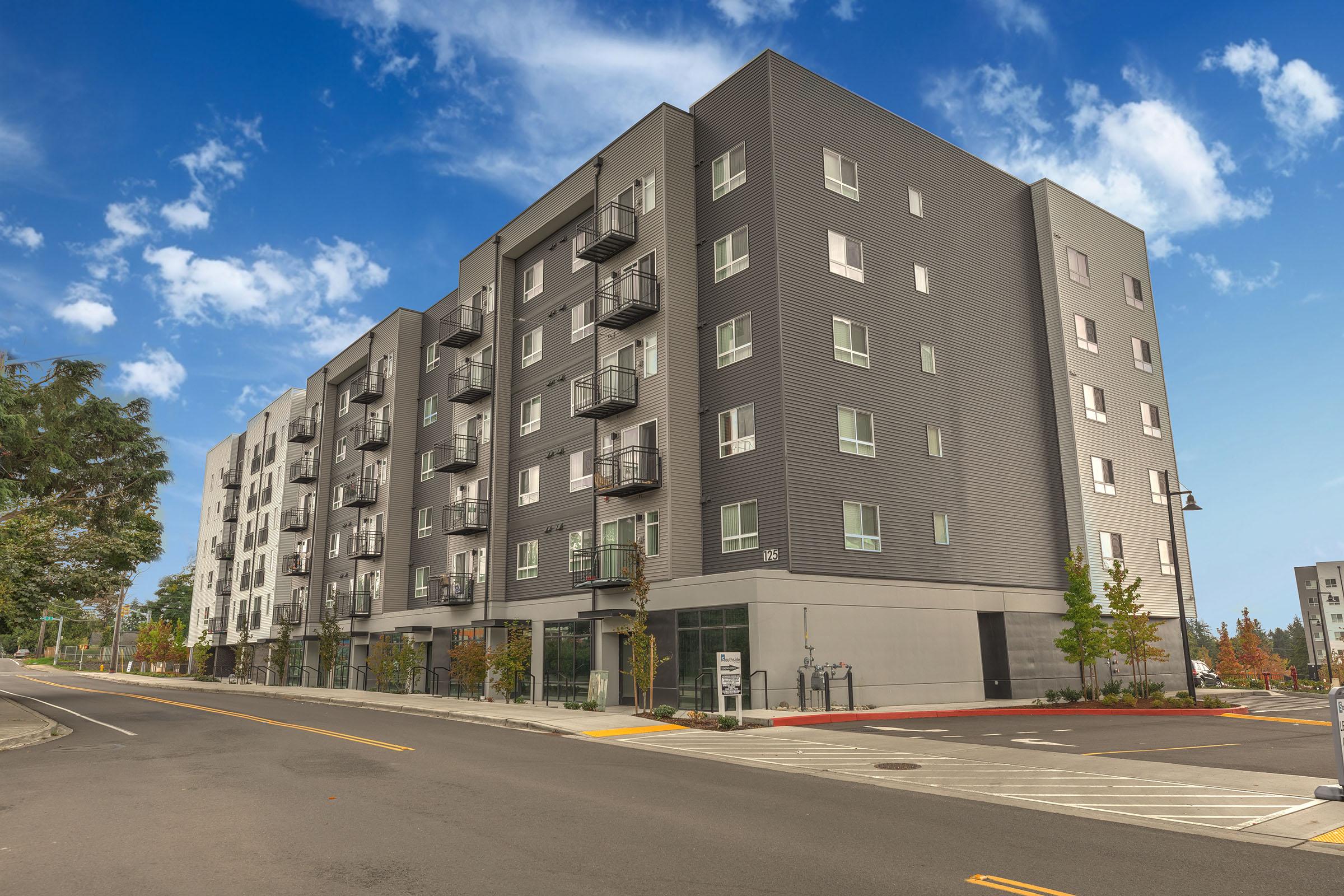 A modern multi-story building with a gray facade, featuring balconies and large windows, situated on a quiet street under a partly cloudy blue sky.