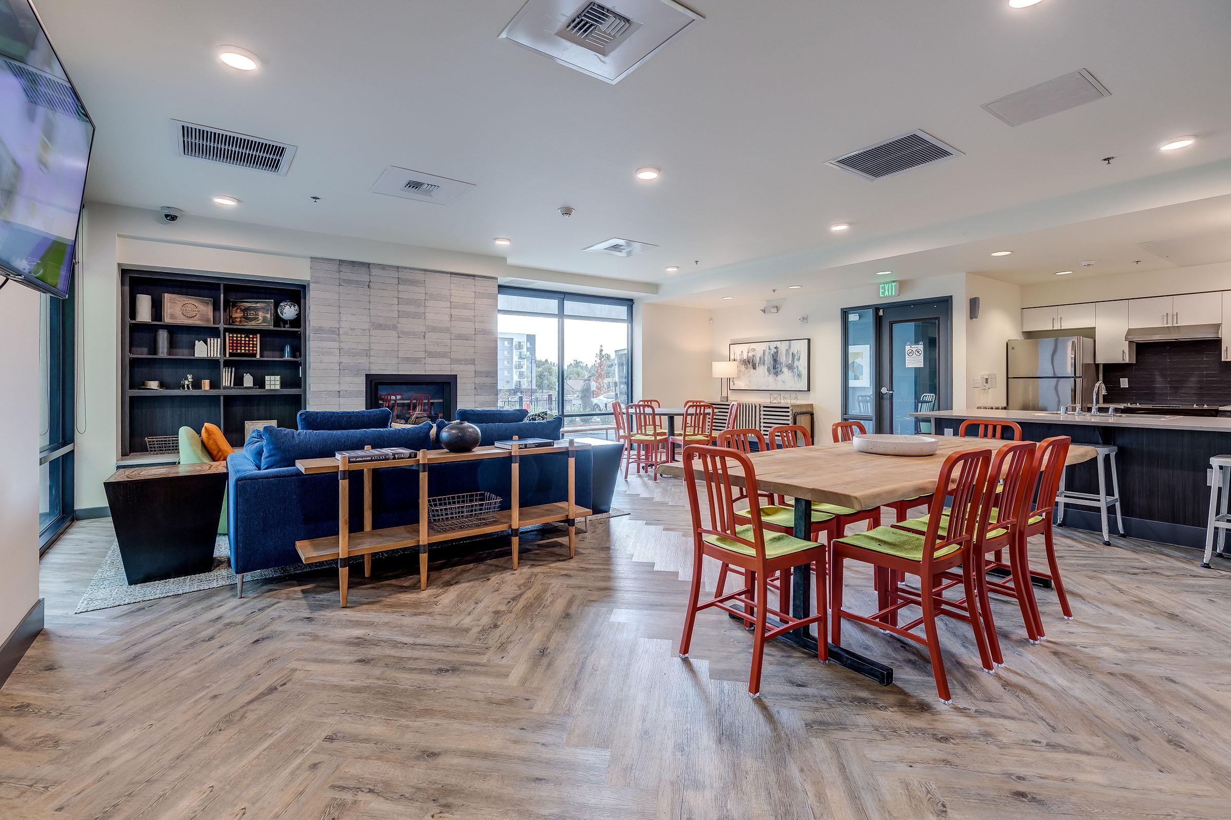 A modern, spacious common area featuring a blue sectional sofa, a wooden table with red chairs, and a kitchenette. Large windows allow natural light to fill the space, with a fireplace and decorative shelves in the background.