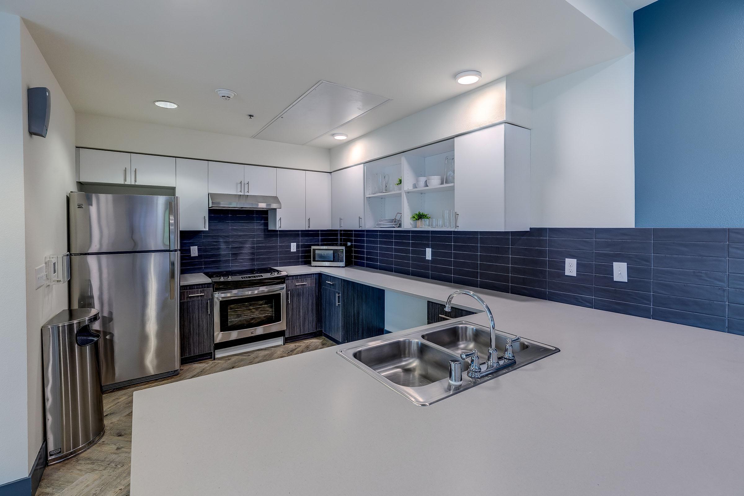 Modern kitchen featuring stainless steel appliances, including a refrigerator and stove, white cabinets, a dark tiled backsplash, and a large countertop with a double sink. The space is well-lit and organized.