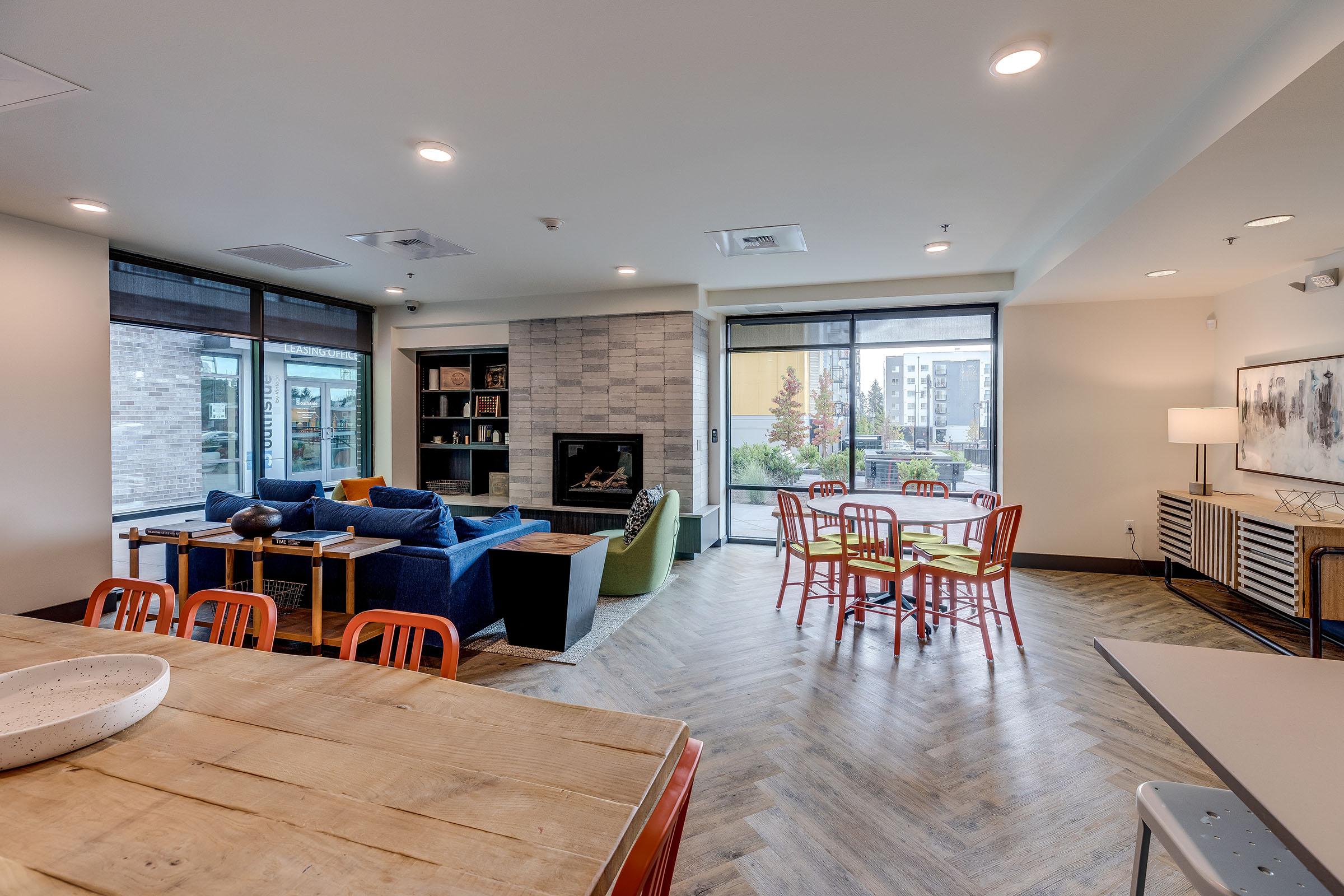 A modern communal living space with a combination of seating areas including a blue couch, a green armchair, and a dining table with red chairs. The room features large windows allowing natural light, a fireplace, and decorative bookshelves. Wood and tile flooring add warmth to the environment.