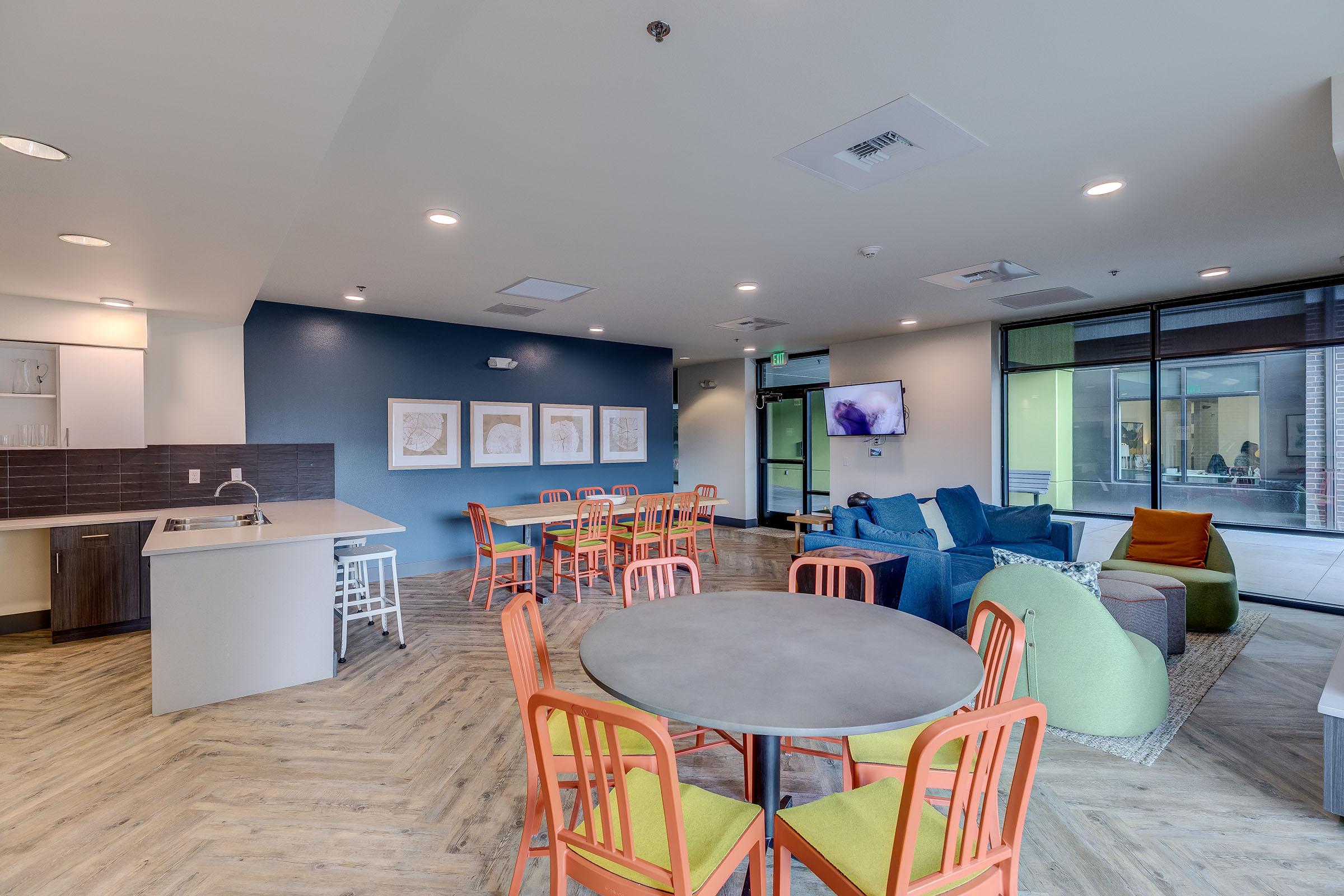 A modern communal living space featuring a kitchenette, multiple dining tables with colorful chairs, and a cozy seating area with sofas and bean bags. Large windows allow natural light to fill the room, and wall art adds a decorative touch.