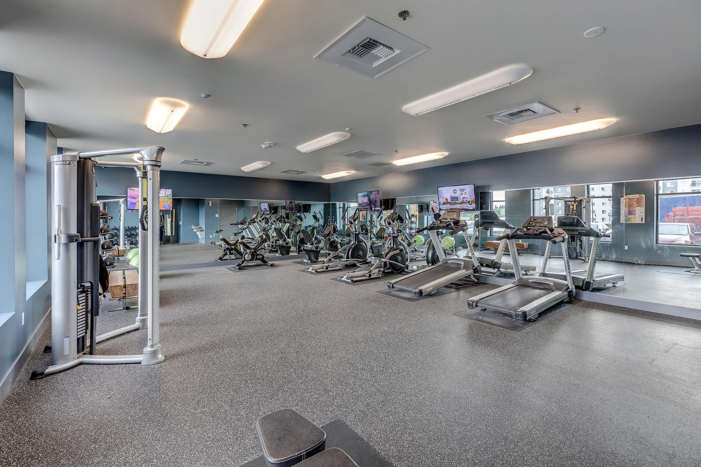 A modern gym interior featuring several exercise machines, including treadmills and stationary bikes, with large mirrors and overhead lights. The space is well-lit and organized, designed for fitness activities.