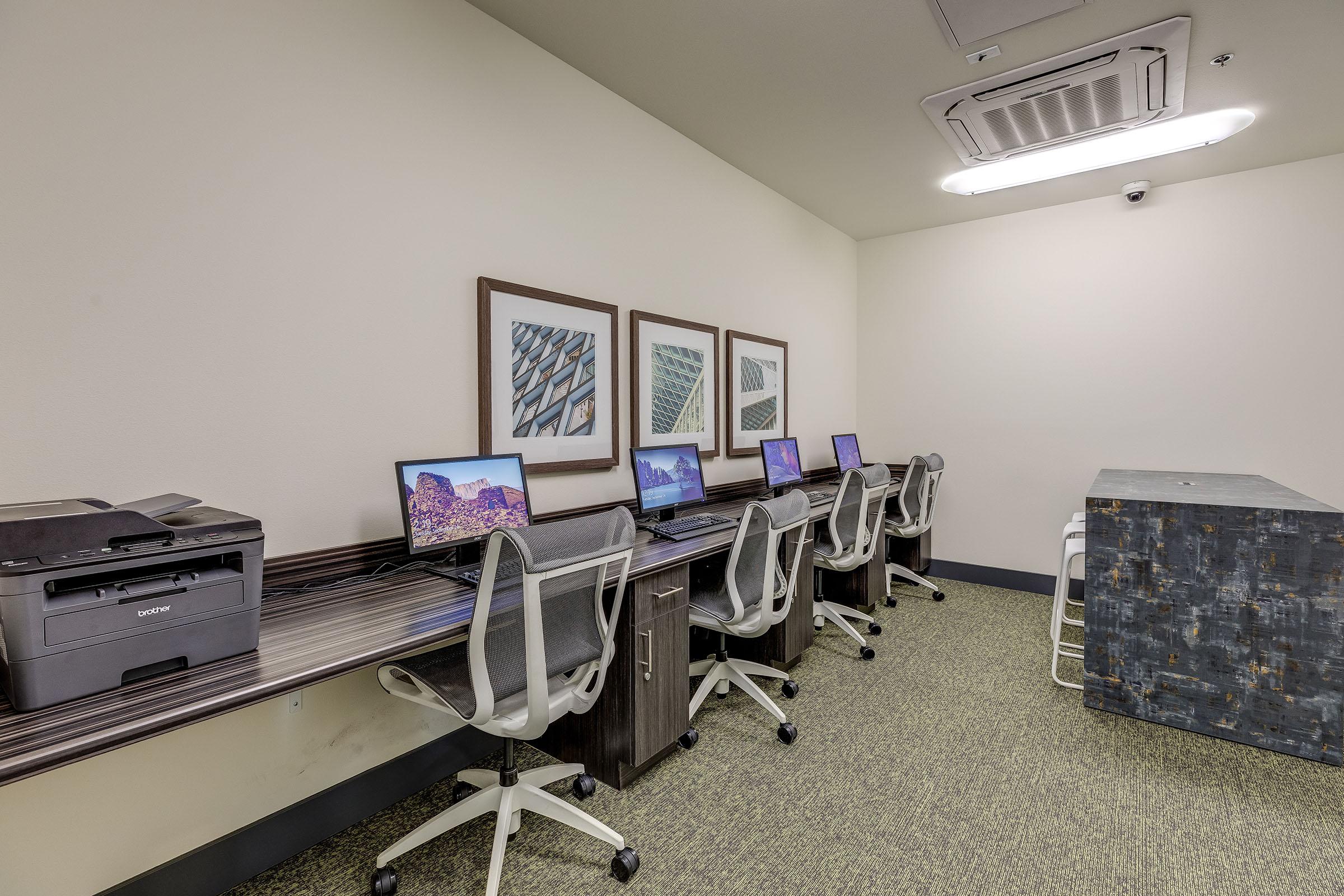 A quiet office space featuring several desktop computers with chairs, a printer, and a small reception or work area. Three framed pictures hang on the wall, and the floor is covered with green carpet.