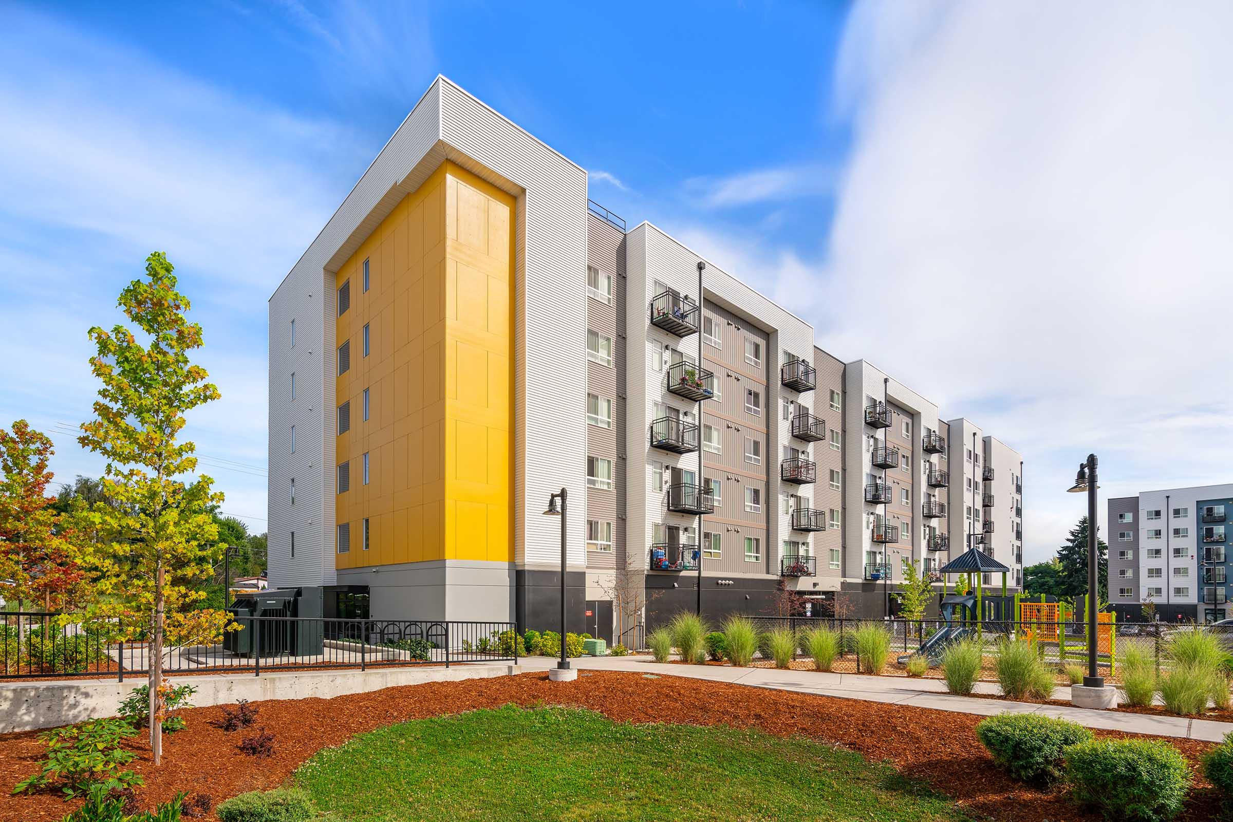 Modern residential apartment building with a yellow accent wall, balconies, and landscaped surroundings featuring trees, shrubs, and pathways, under a bright blue sky.