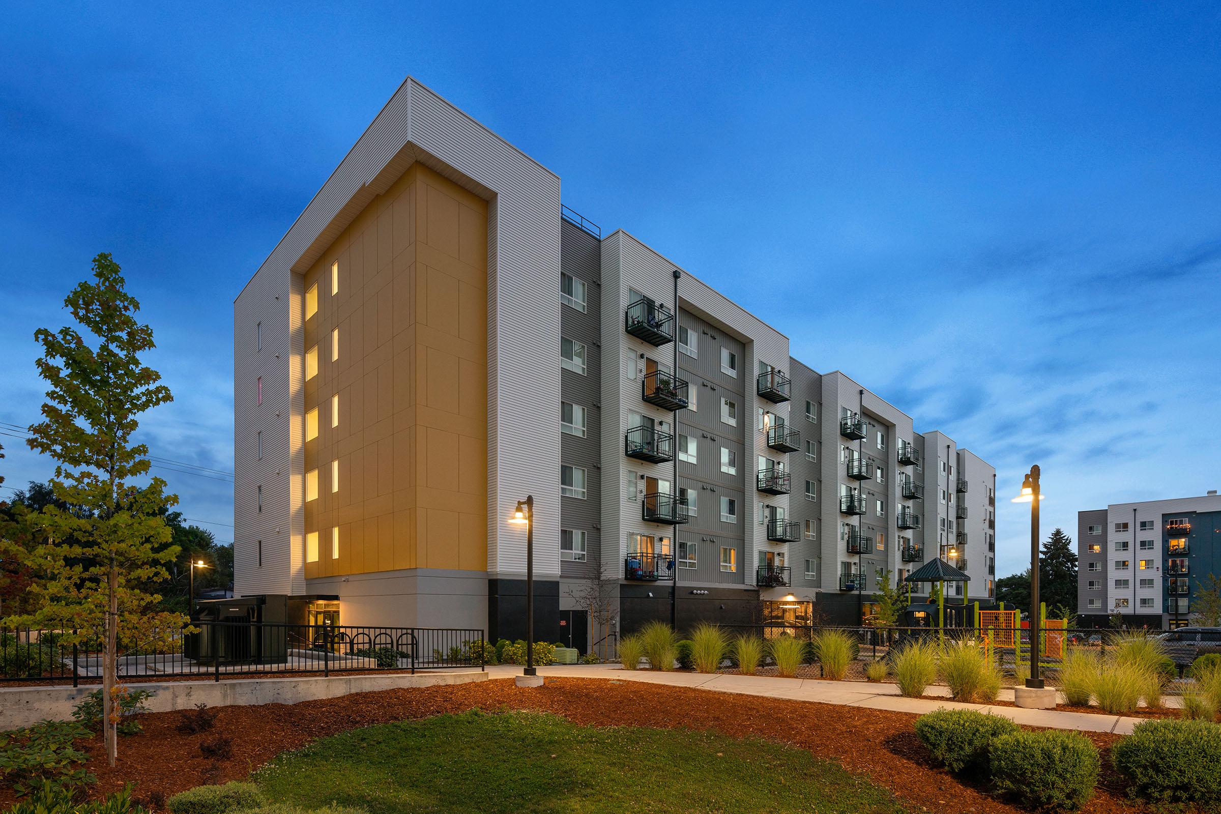 Modern apartment building with a sleek design, featuring multiple balconies, landscaped green space, and outdoor lighting, set against a twilight sky.